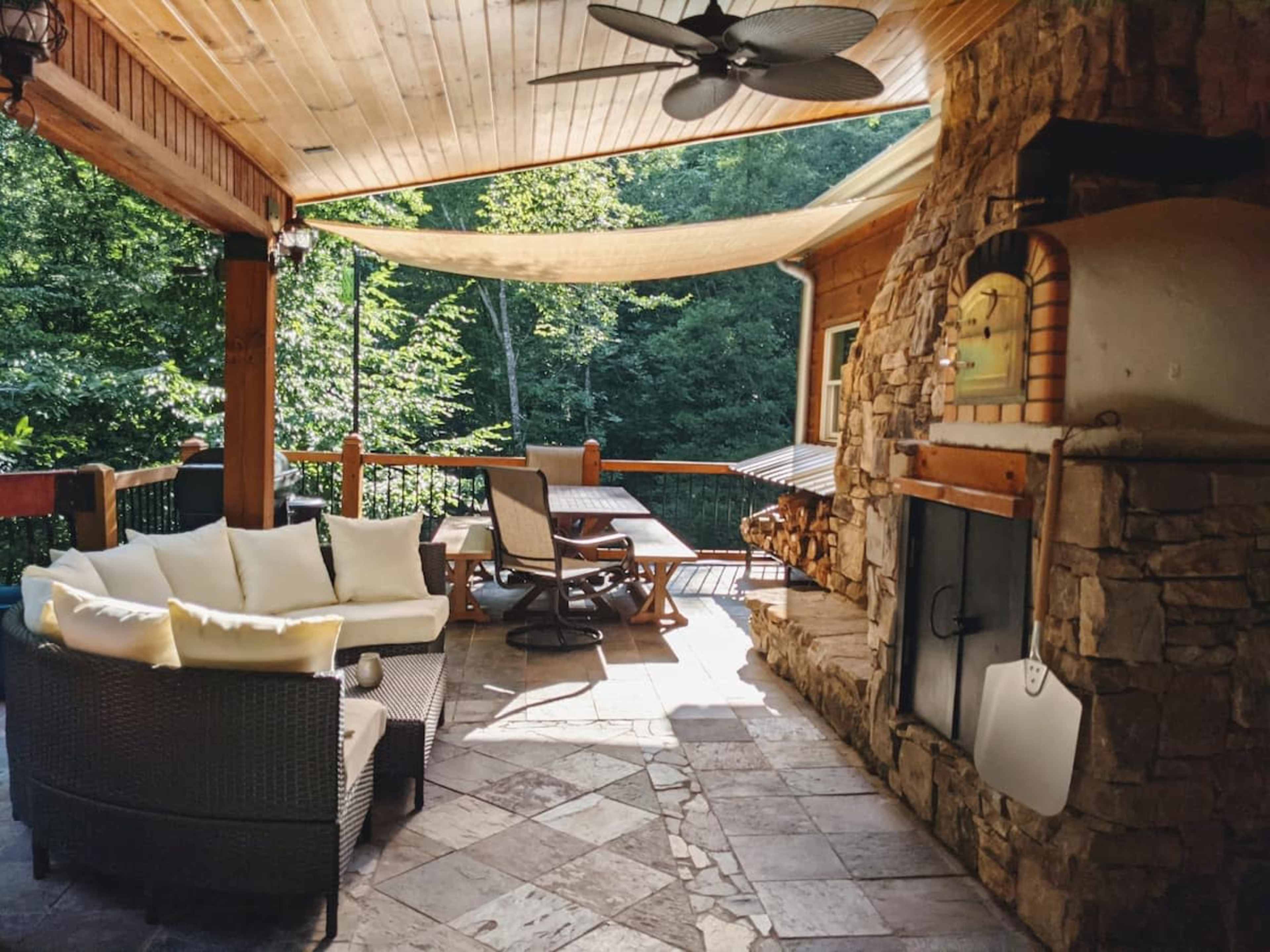 A covered outdoor patio featuring a stone fireplace, a dining table, and a cushioned seating arrangement, surrounded by greenery.