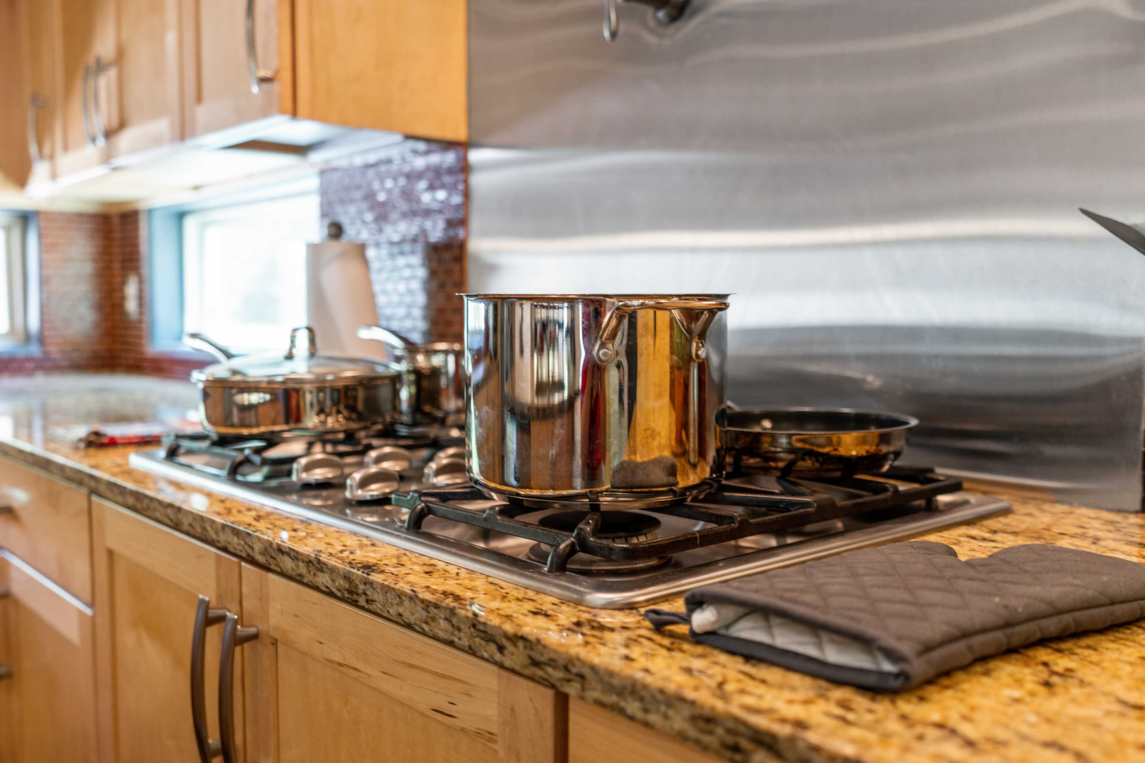 A stovetop with several pots and pans is set against a backdrop of wooden cabinetry and a stainless steel backsplash.