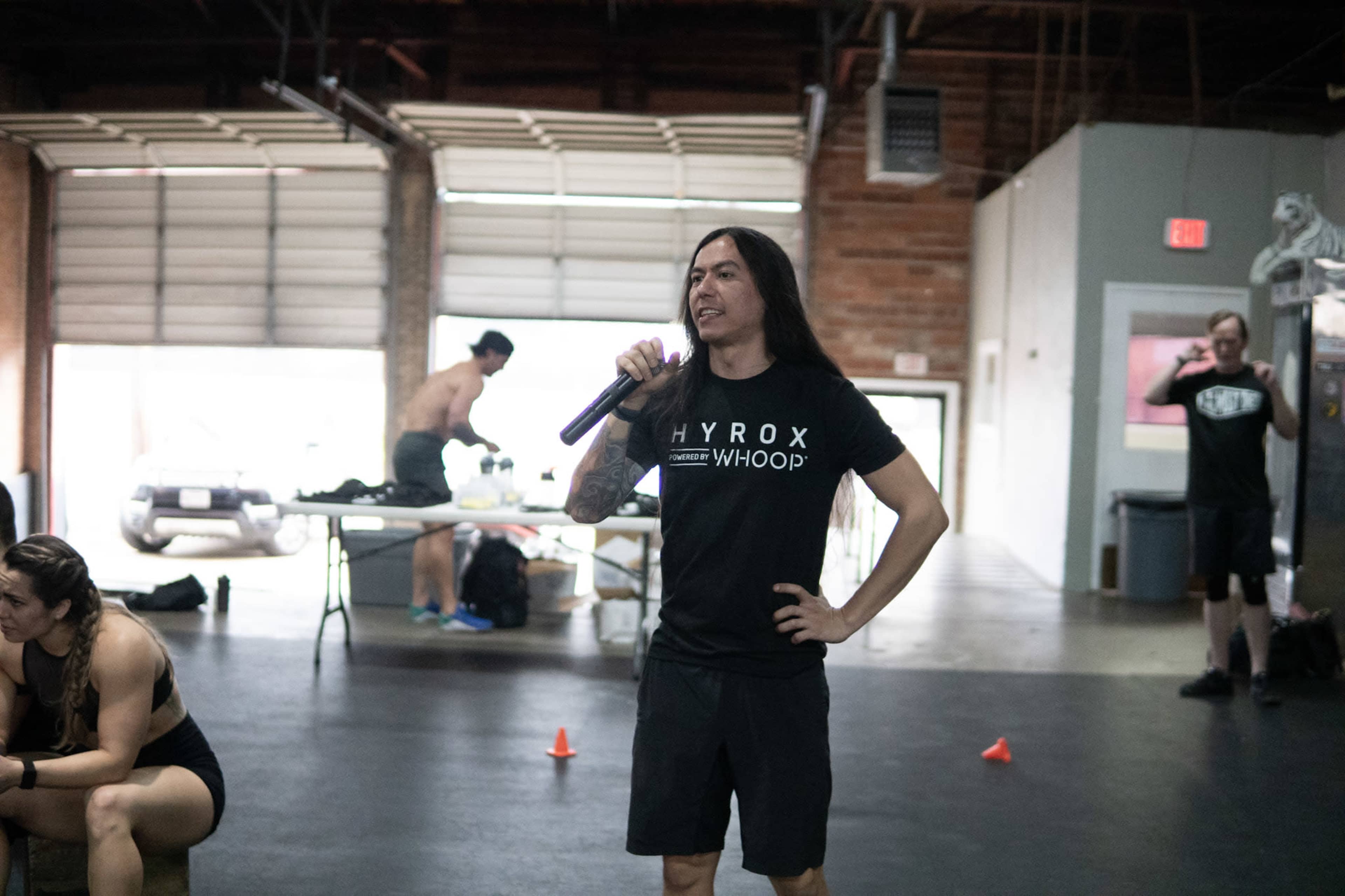 A person in a black t-shirt stands in a gym, holding a microphone while others train in the background.