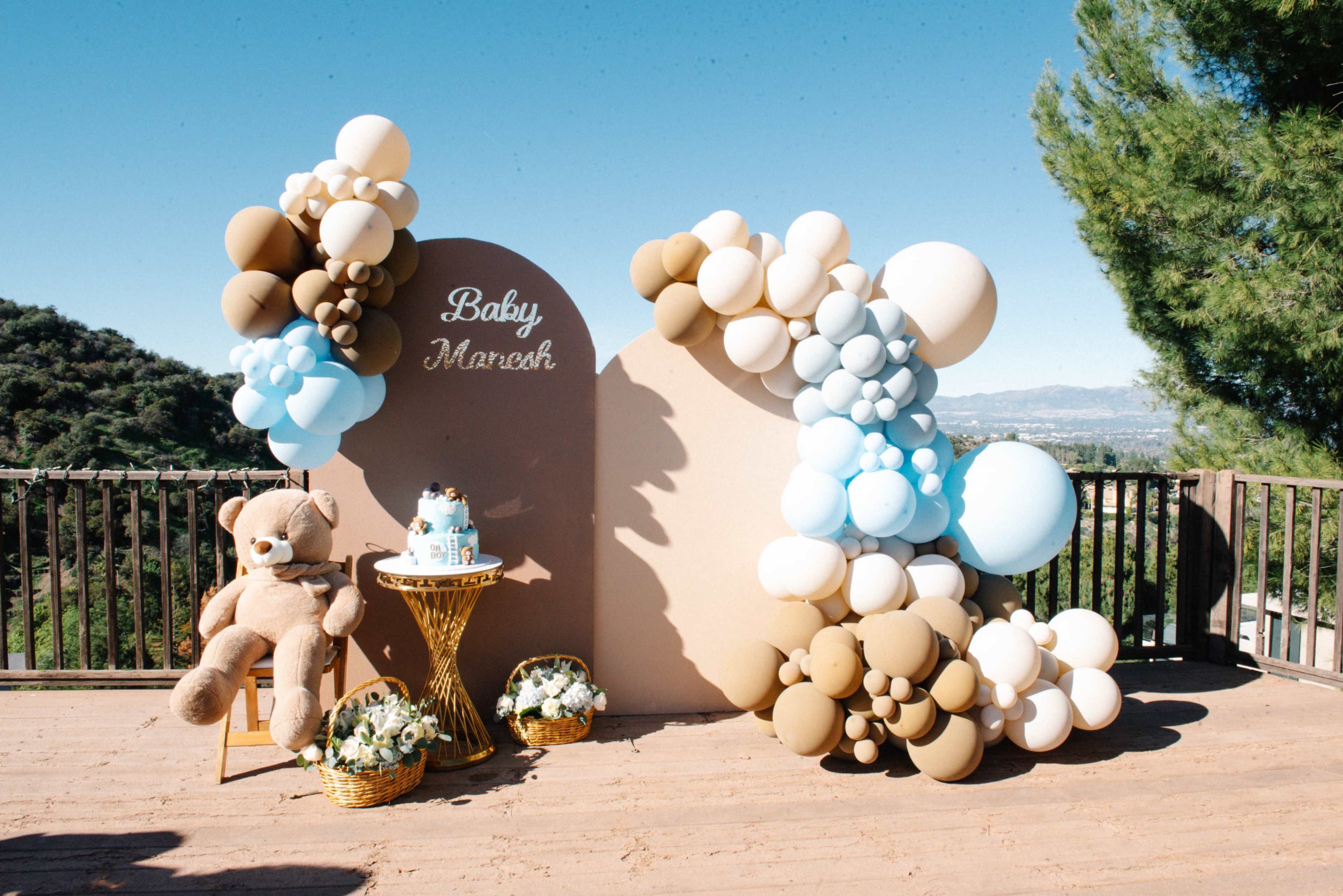 The scene features a decorative display for a baby-themed event, with a backdrop of balloons in pastel colors, a plush teddy bear, and floral arrangements on a wooden deck overlooking a mountainous landscape.