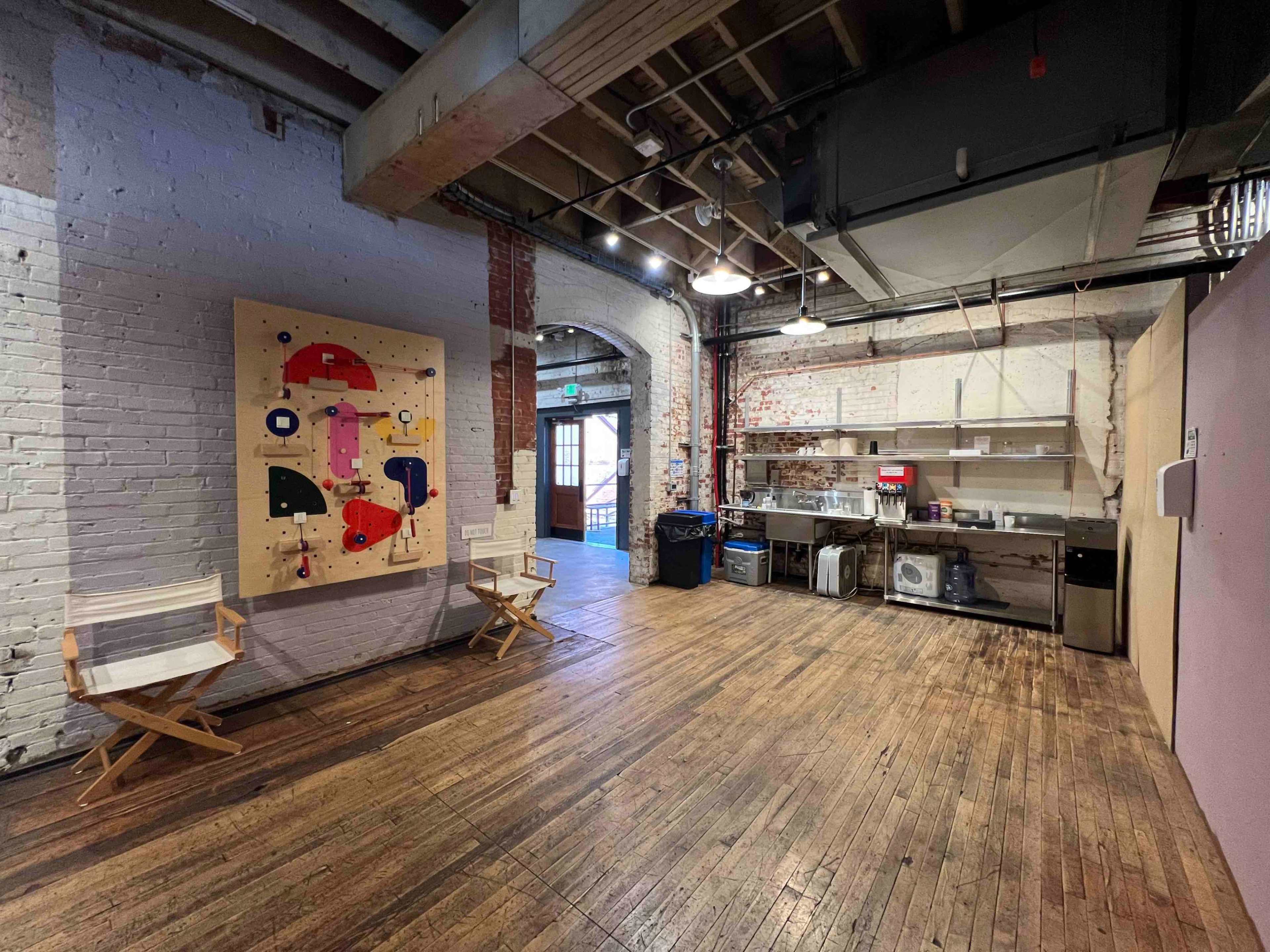 A spacious room with wooden floors, featuring a colorful abstract painting on the wall and a kitchen area with appliances in the background.
