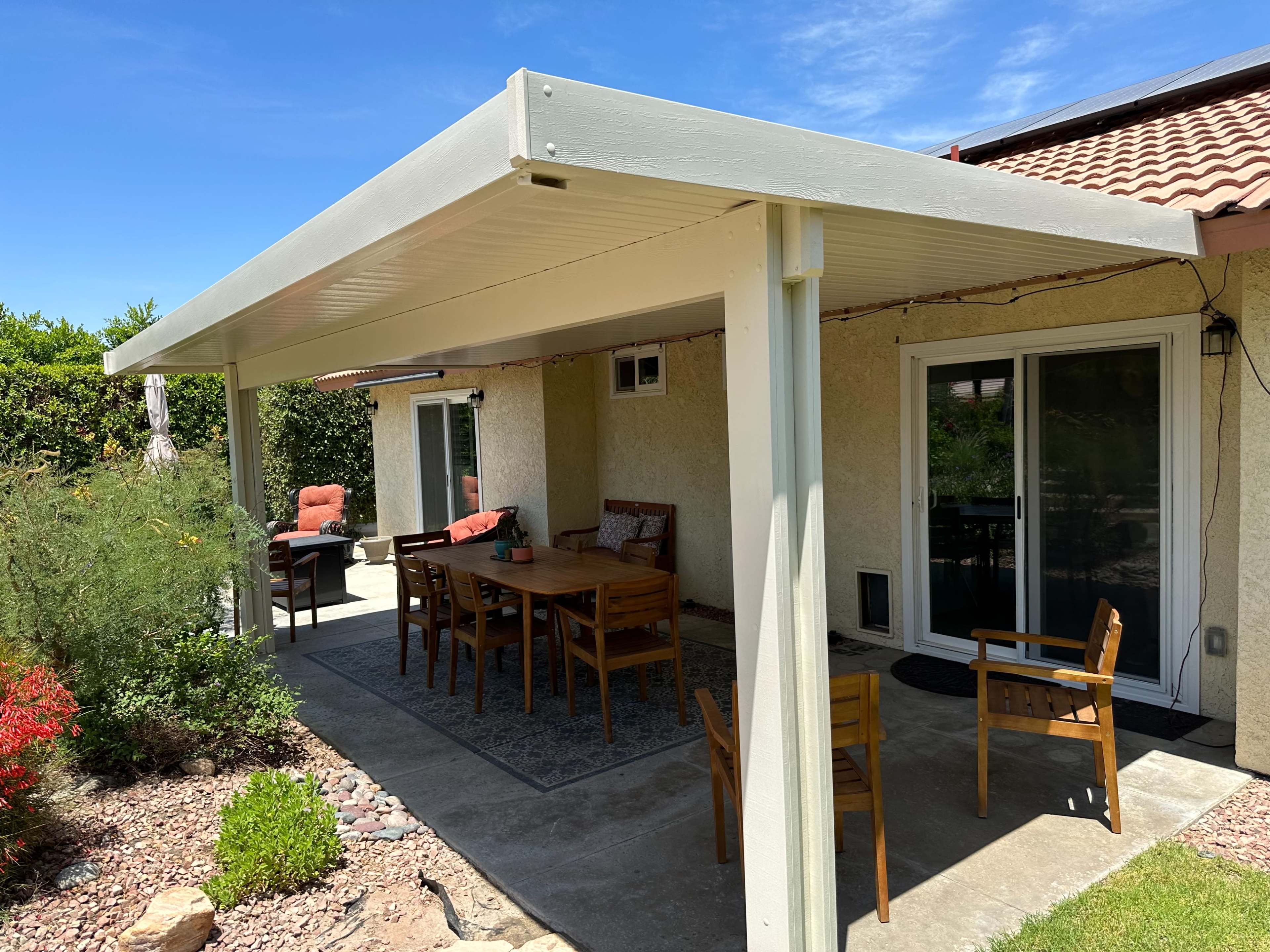 The image shows a covered patio area with a wooden dining table and chairs, surrounded by lush greenery.
