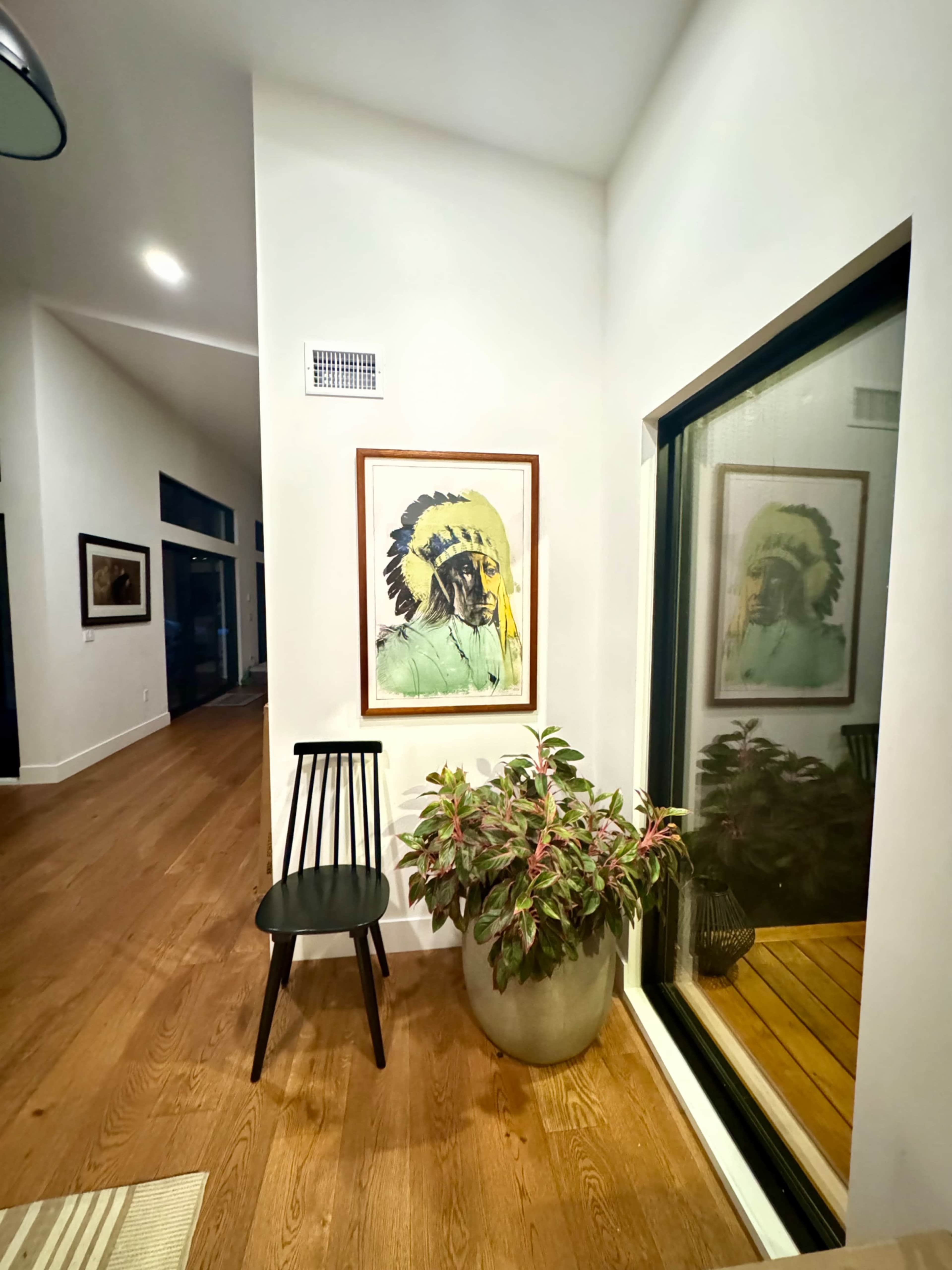 A chair sits beside a large potted plant in a corner of a room, featuring a window that reflects a framed artwork of a person wearing a headdress.
