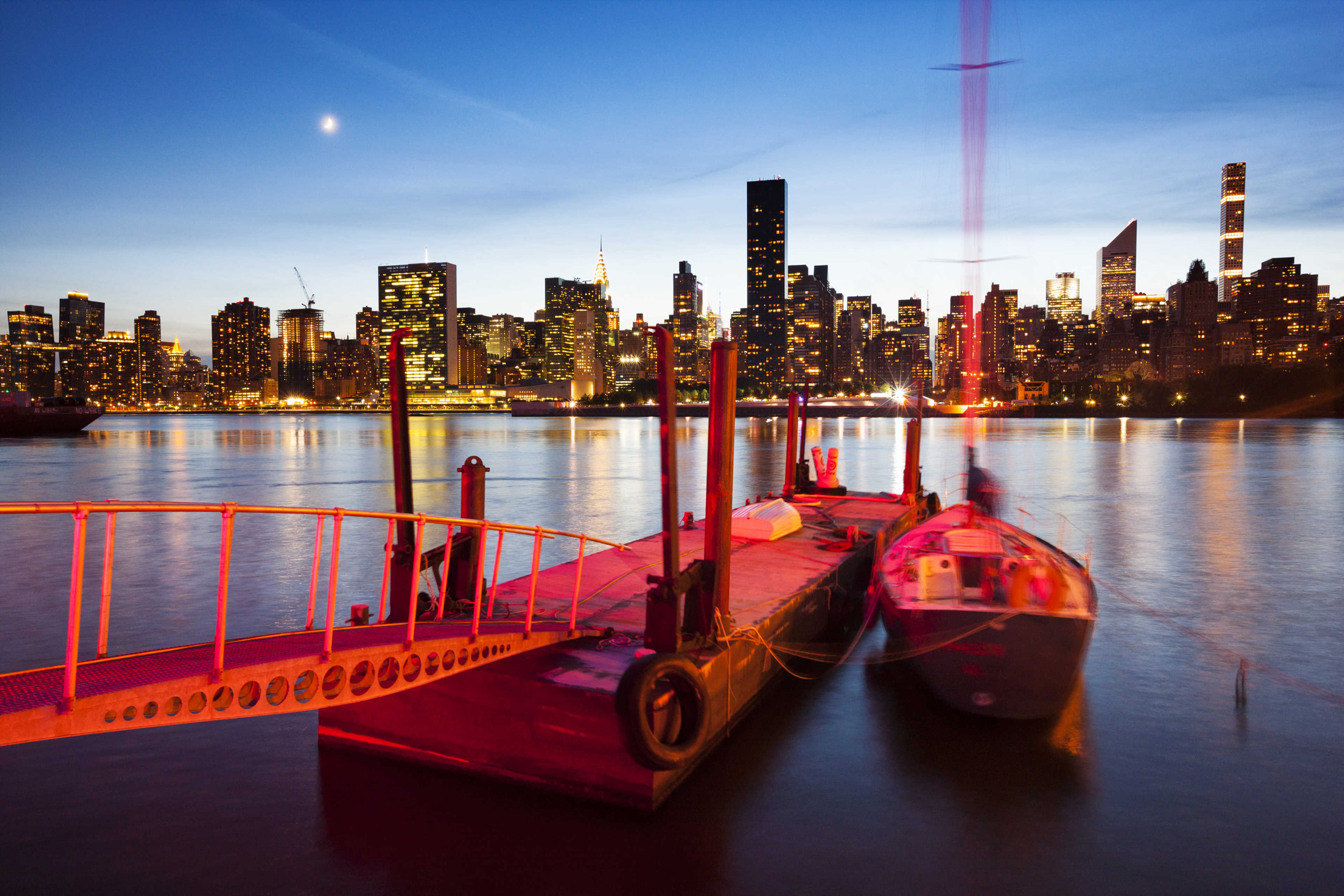 A boat is docked at a pier with the skyline of New York City lit up at dusk.