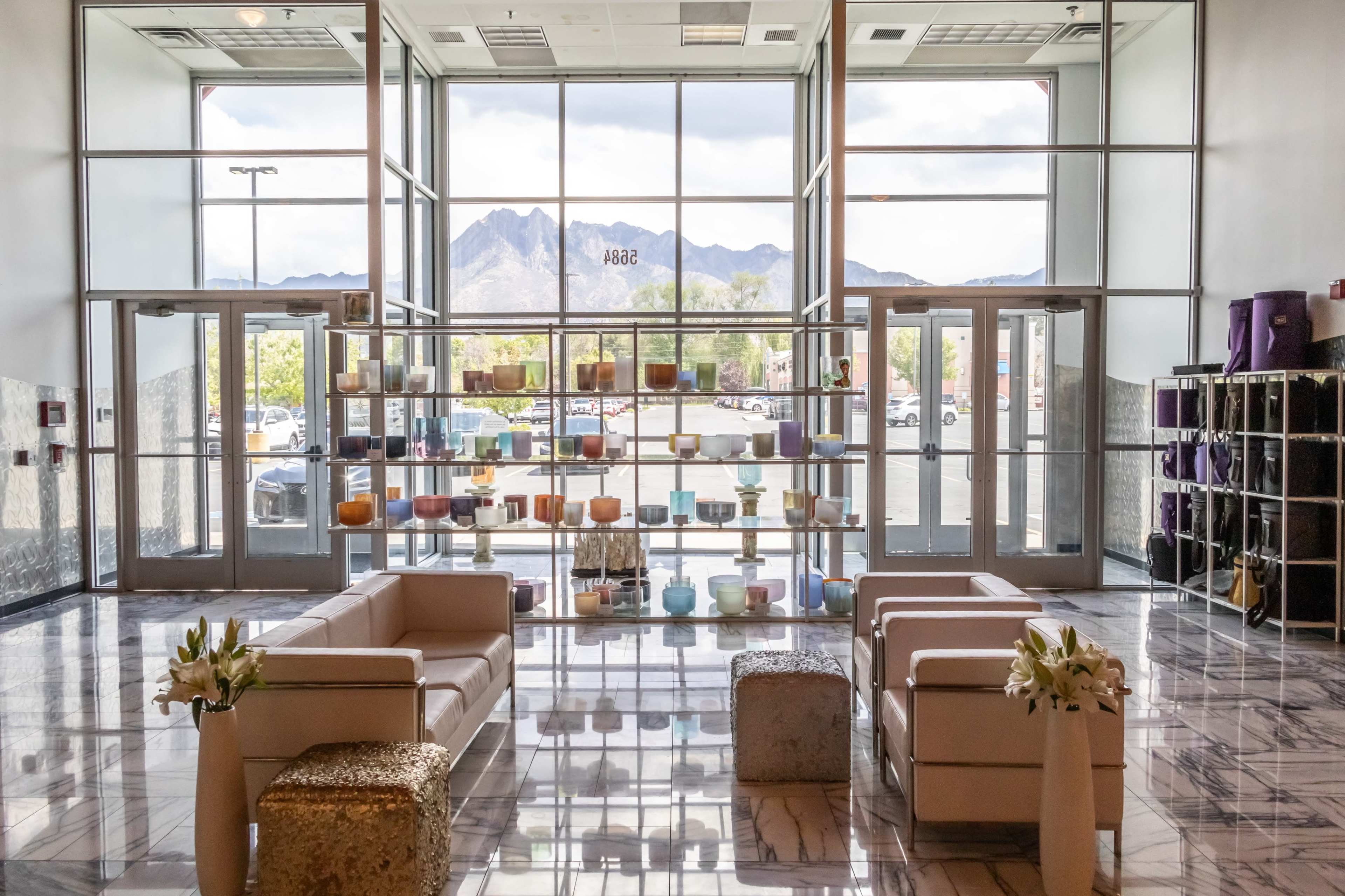 The image shows a modern lobby with two white couches, a display shelf filled with candles, and large windows offering a view of distant mountains.