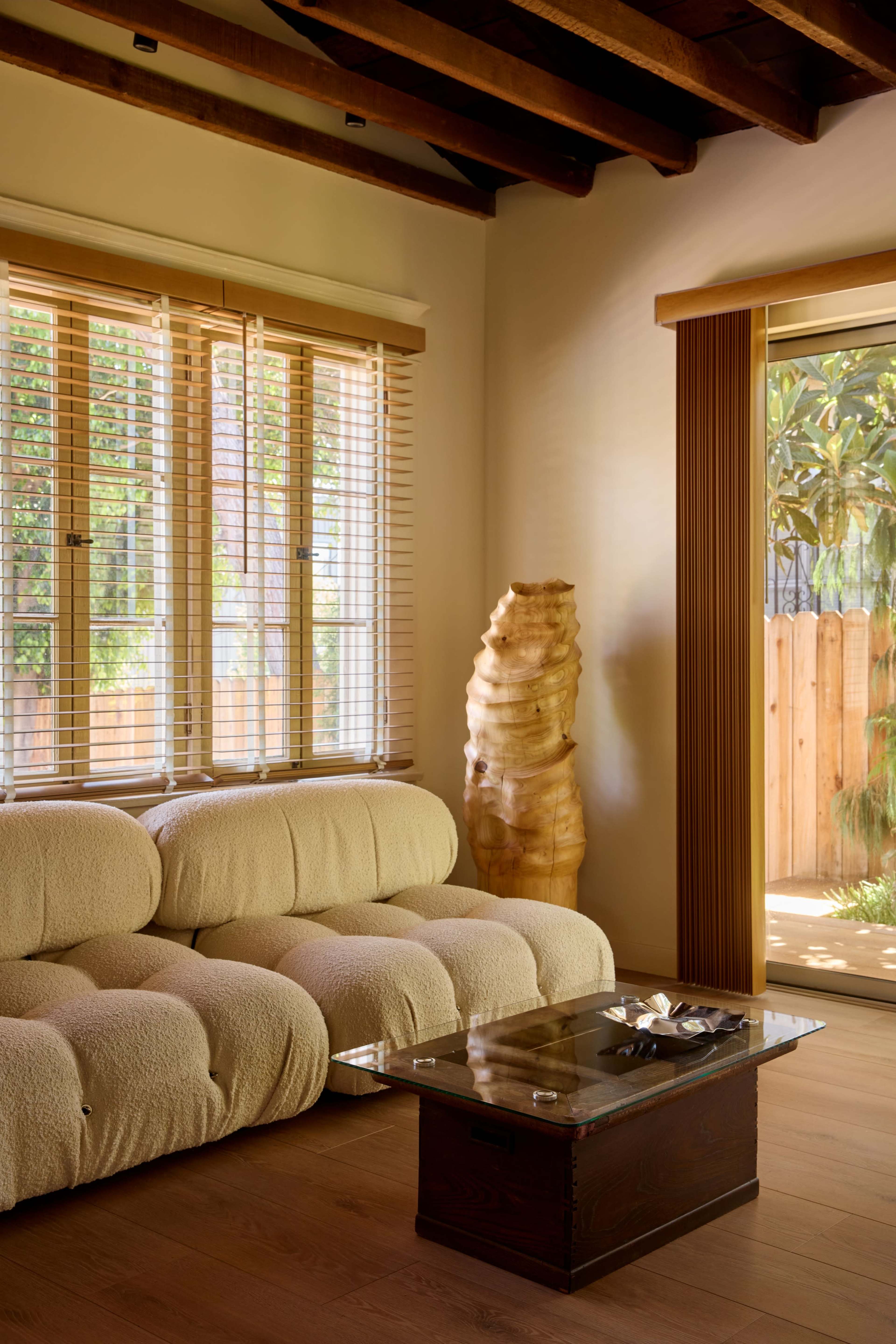 The image shows a cozy living room featuring a beige sectional sofa, a glass coffee table, and a wooden sculpture beside large windows.
