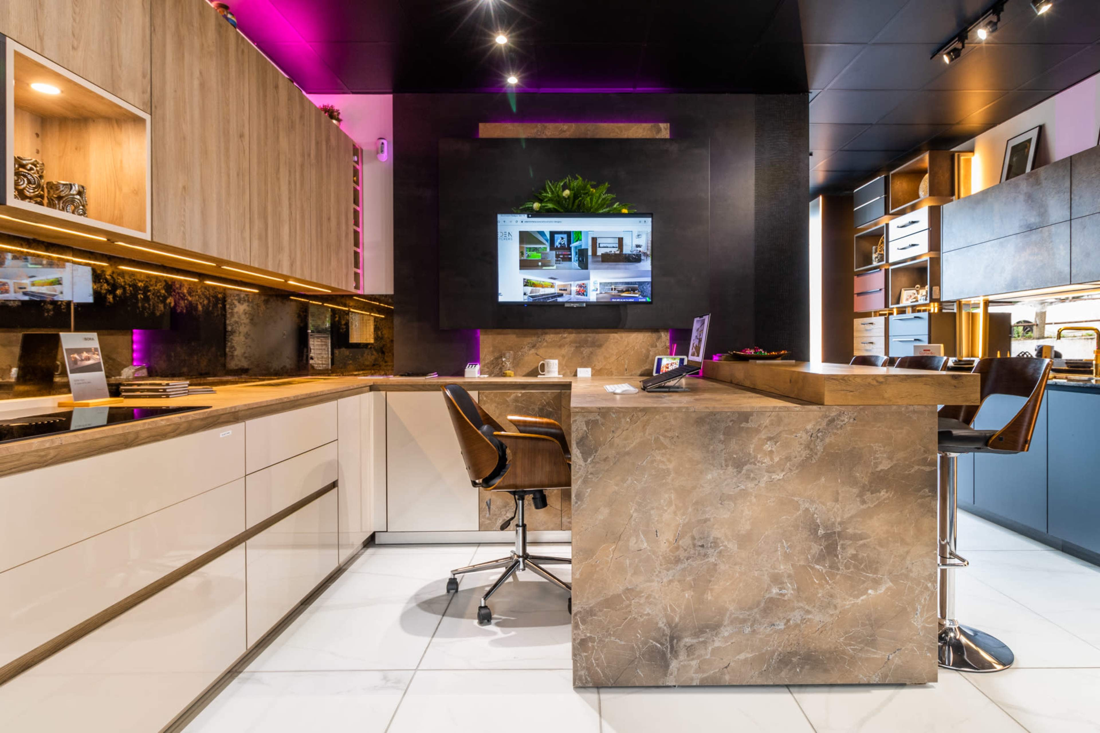 A modern kitchen display features sleek cabinetry, a large stone-topped island, and a digital screen mounted on the wall.