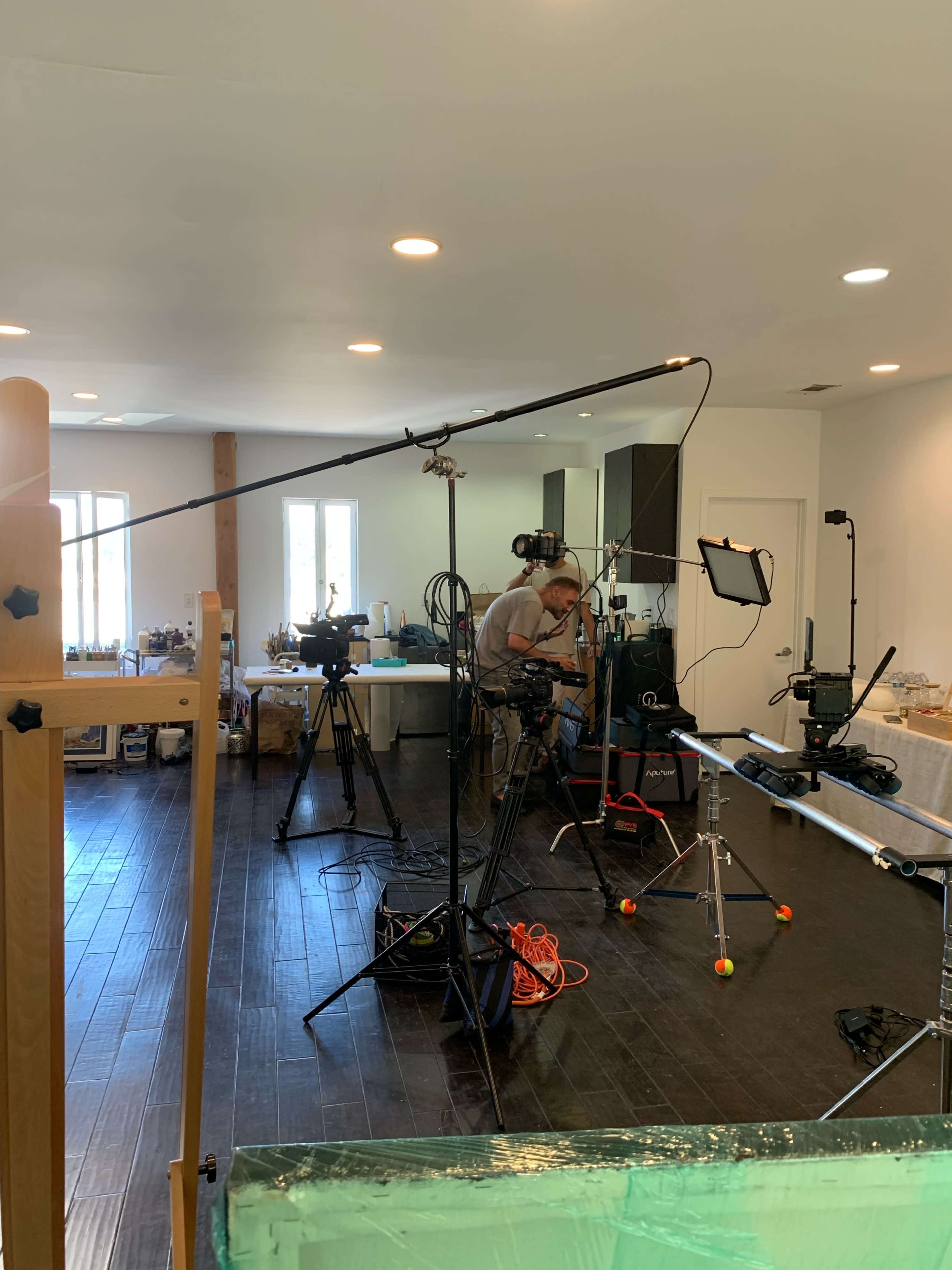 A film production setup is staged in a room with multiple cameras, lighting equipment, and a microphone on a boom.