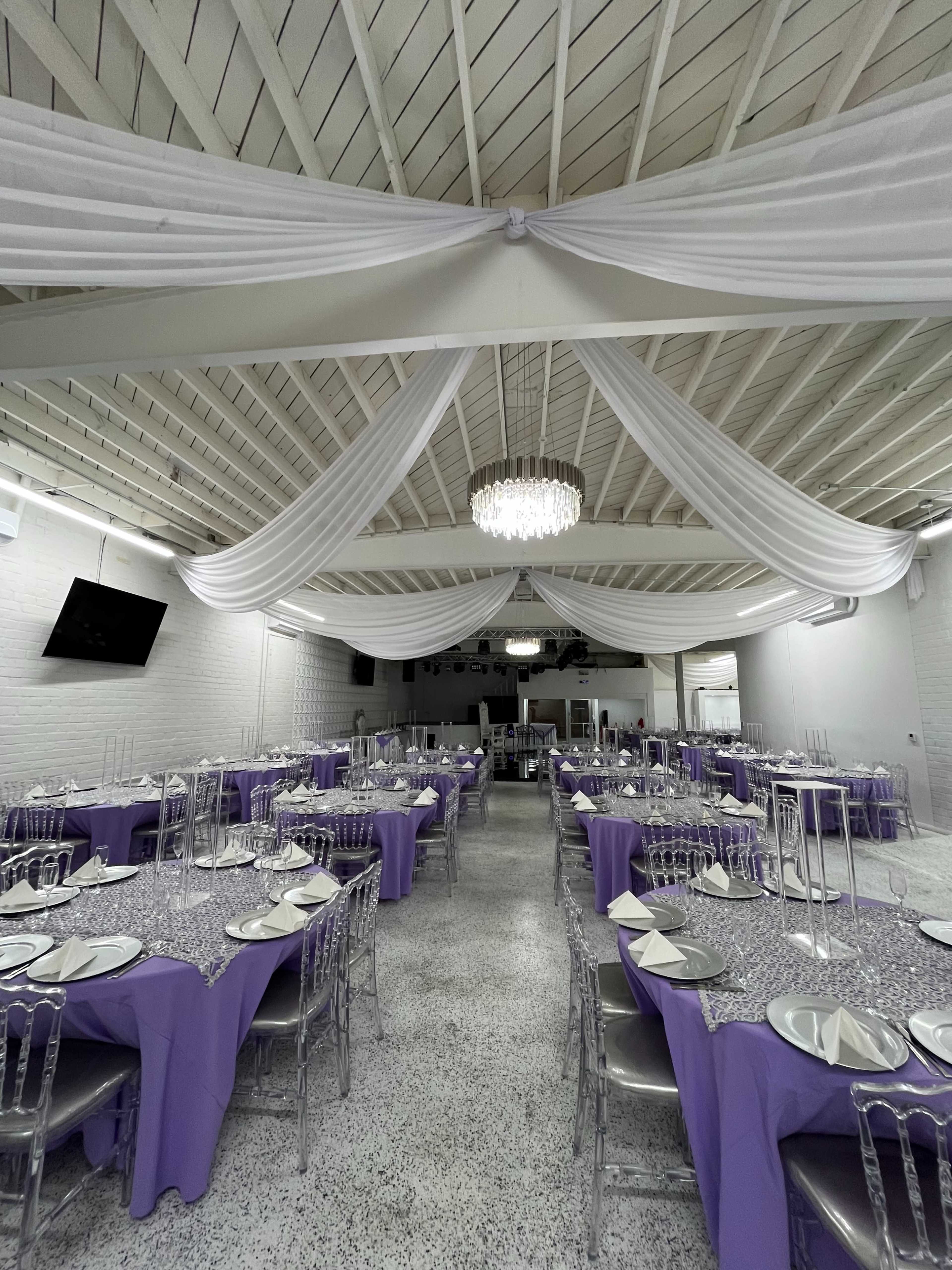 The image shows a large event space set up for a formal gathering, featuring several tables covered with purple tablecloths and adorned with elegant place settings under a draped ceiling and a chandelier.