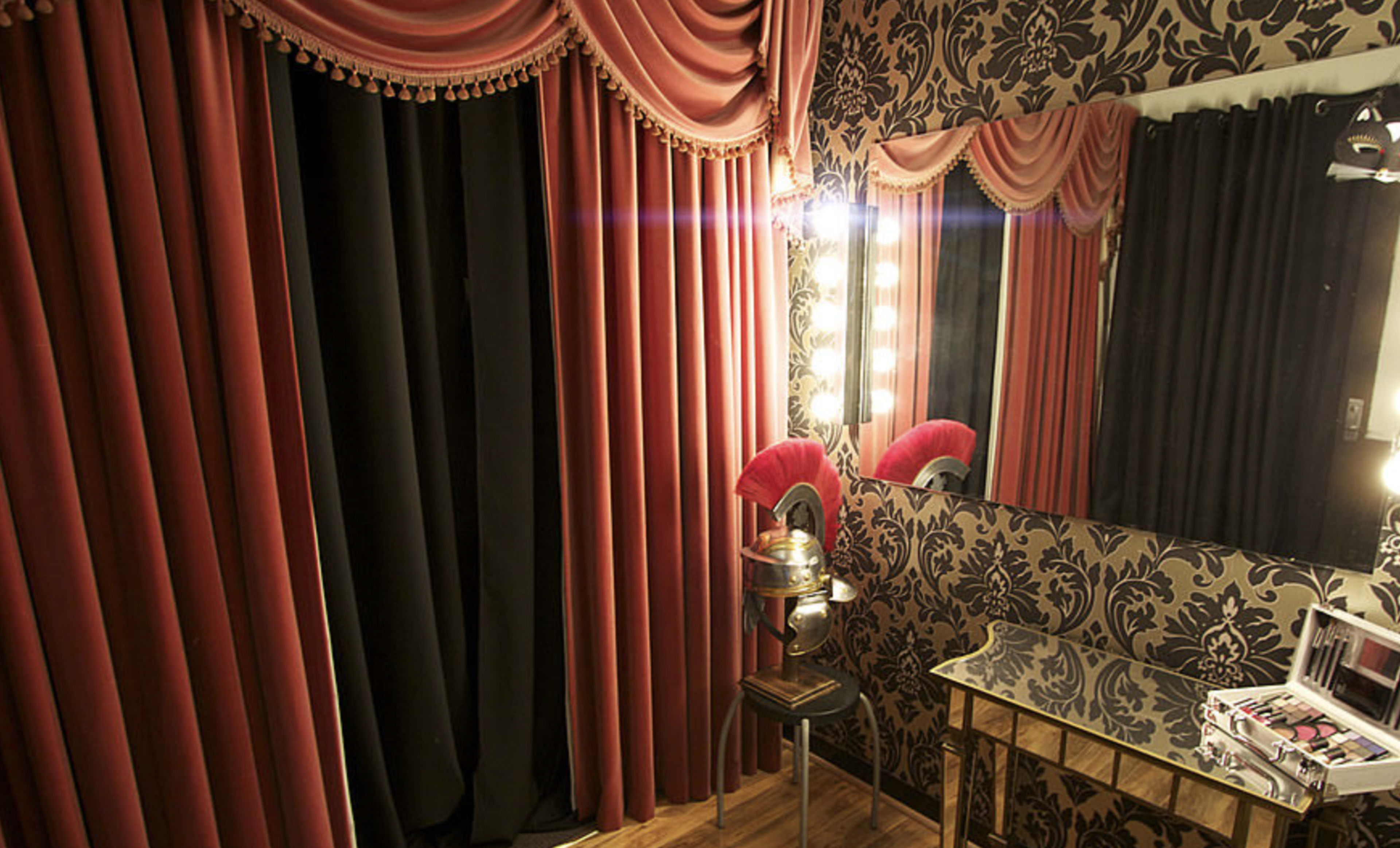 A dressing room with red curtains, a large mirror surrounded by lights, and a vanity table with cosmetic products.