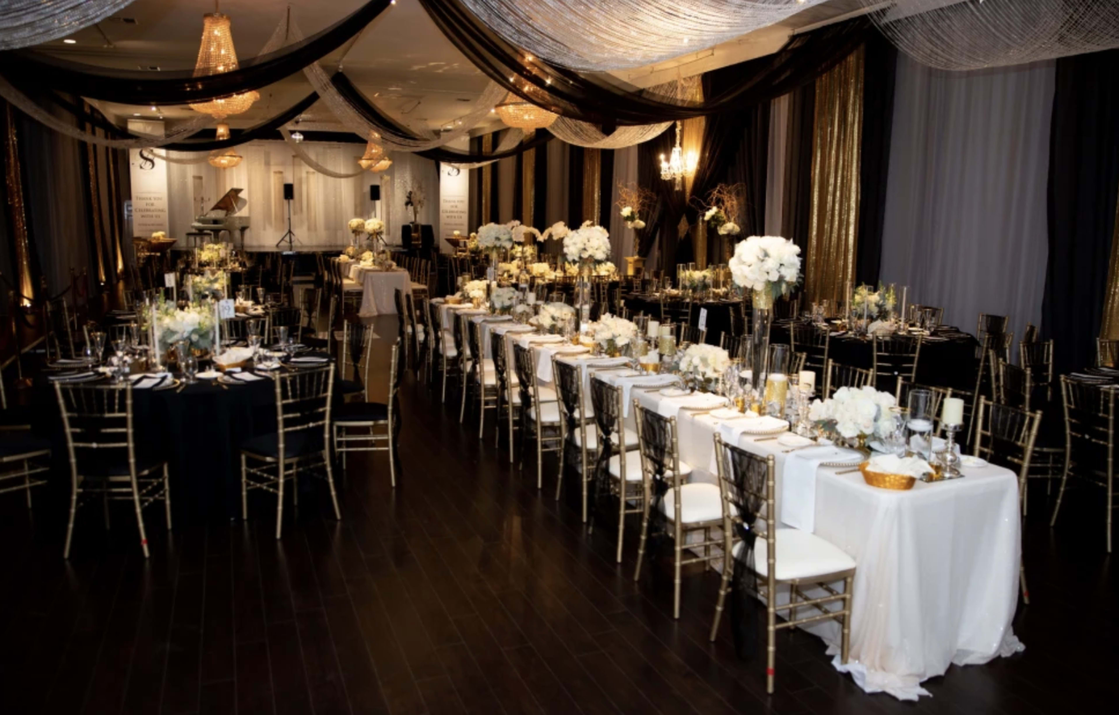 An elegantly arranged banquet hall with long tables set for a formal event, decorated with white flowers and black and gold drapery.