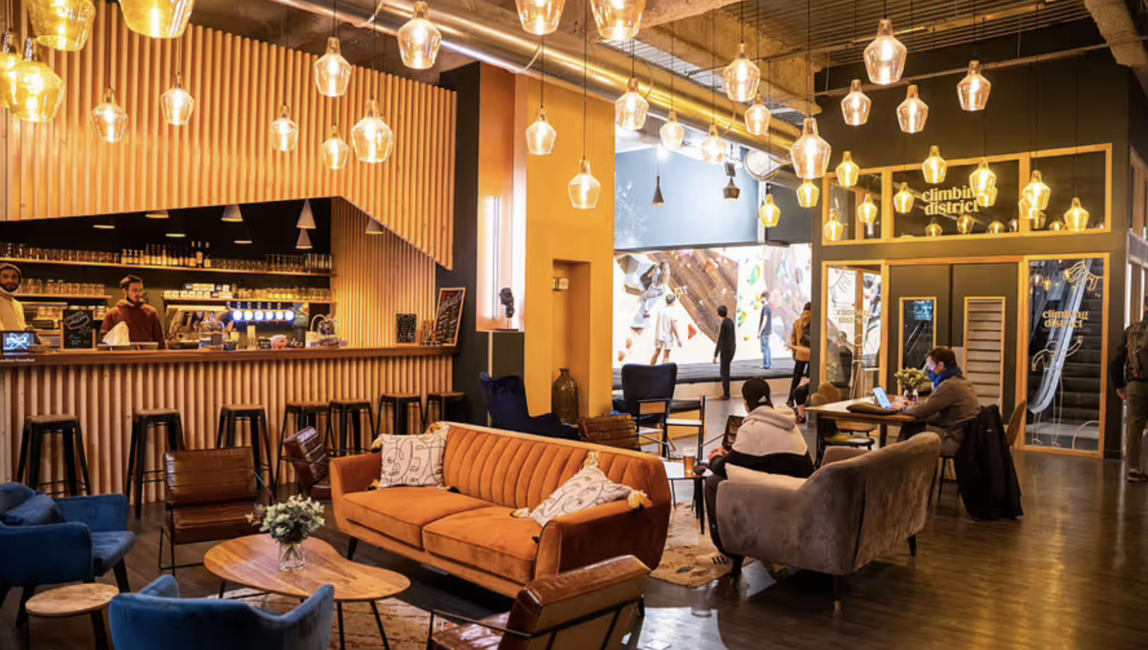 A vibrant indoor climbing gym with a café area featuring a wooden bar, a seating area with sofas, and climbing walls visible through large windows.