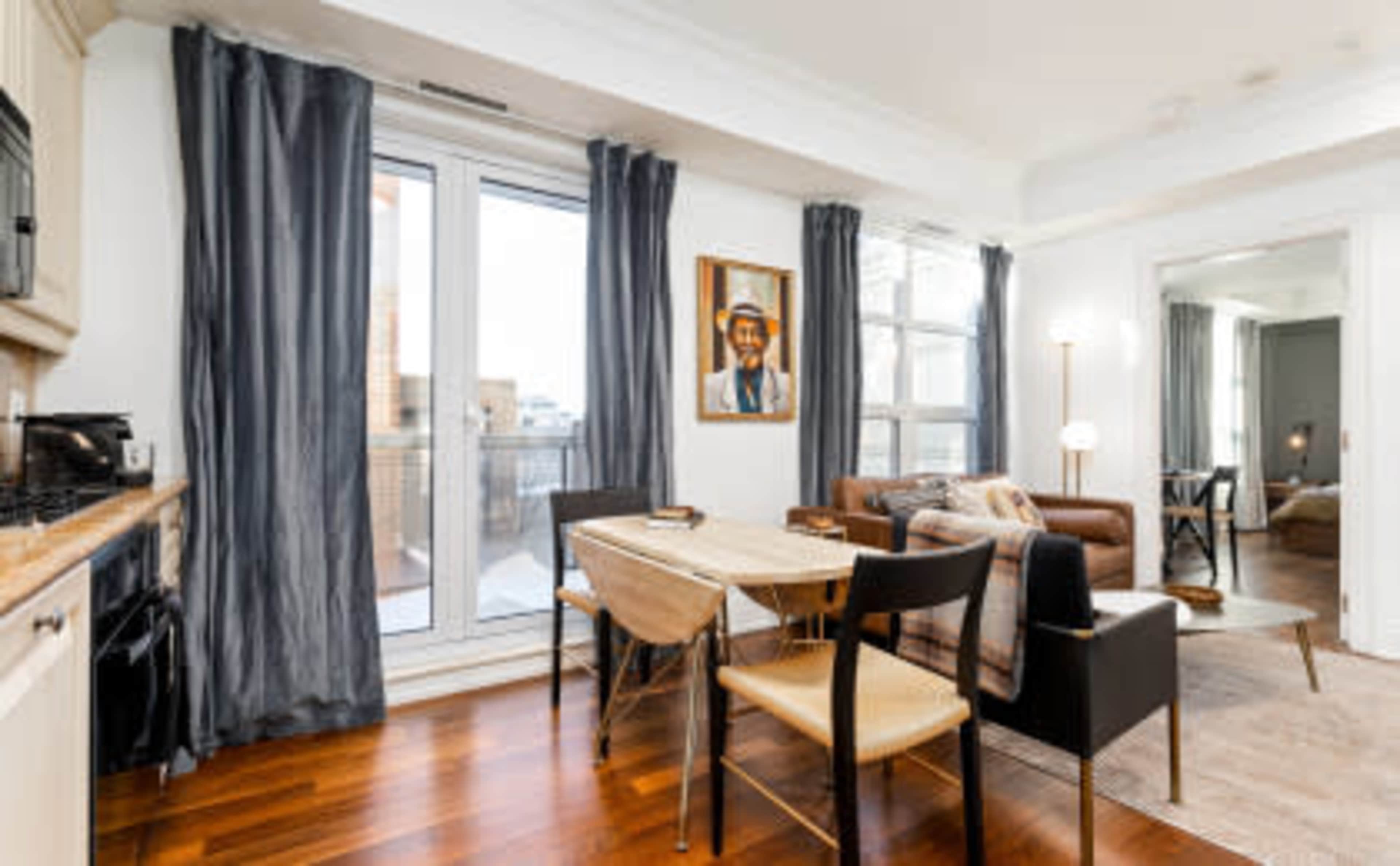 A modern apartment features a dining area with a wooden table and chairs, large windows with gray curtains, and a living area with a leather couch and a portrait on the wall.