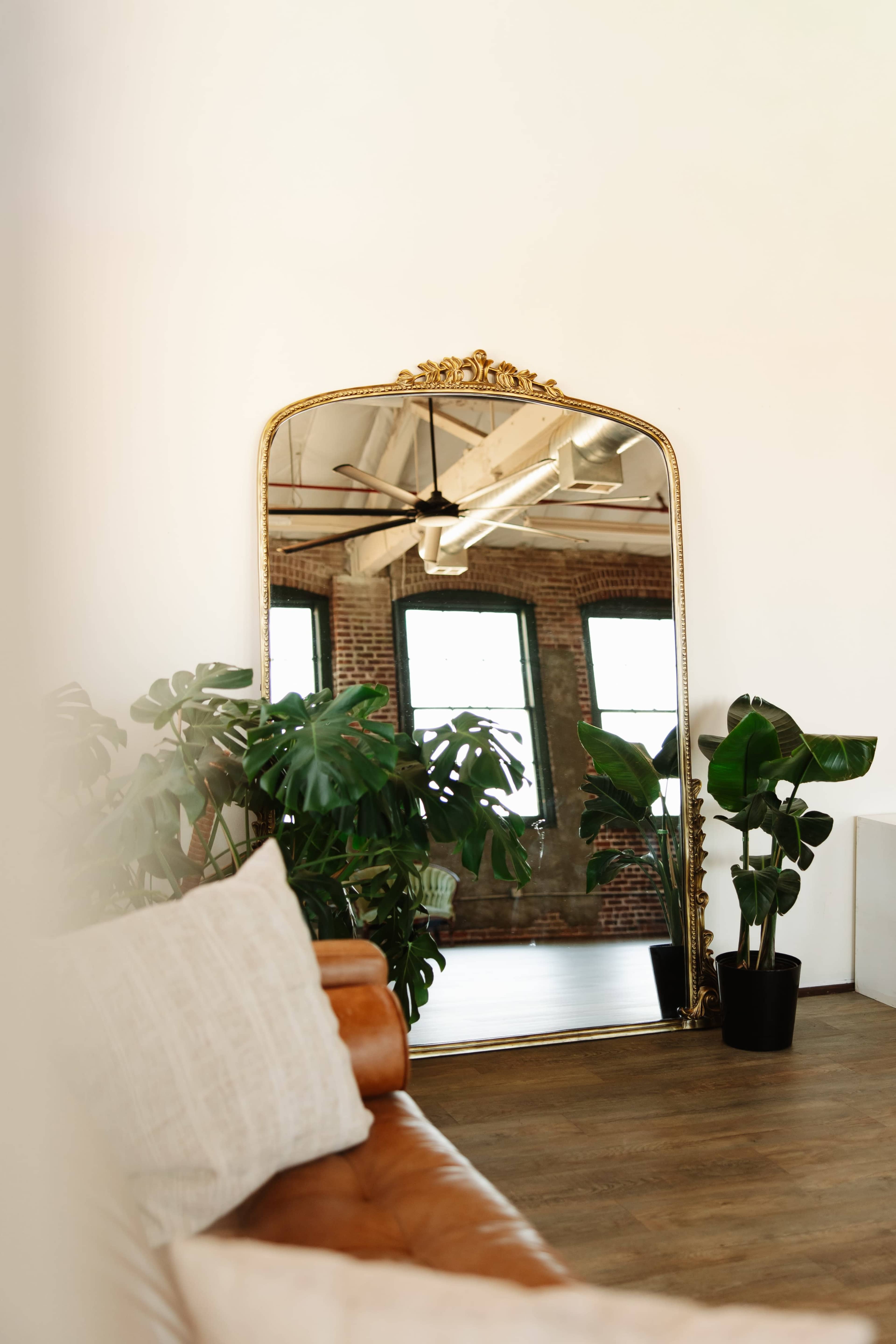 A large ornate mirror reflects a room with industrial-style windows and potted plants beside a brown leather sofa.