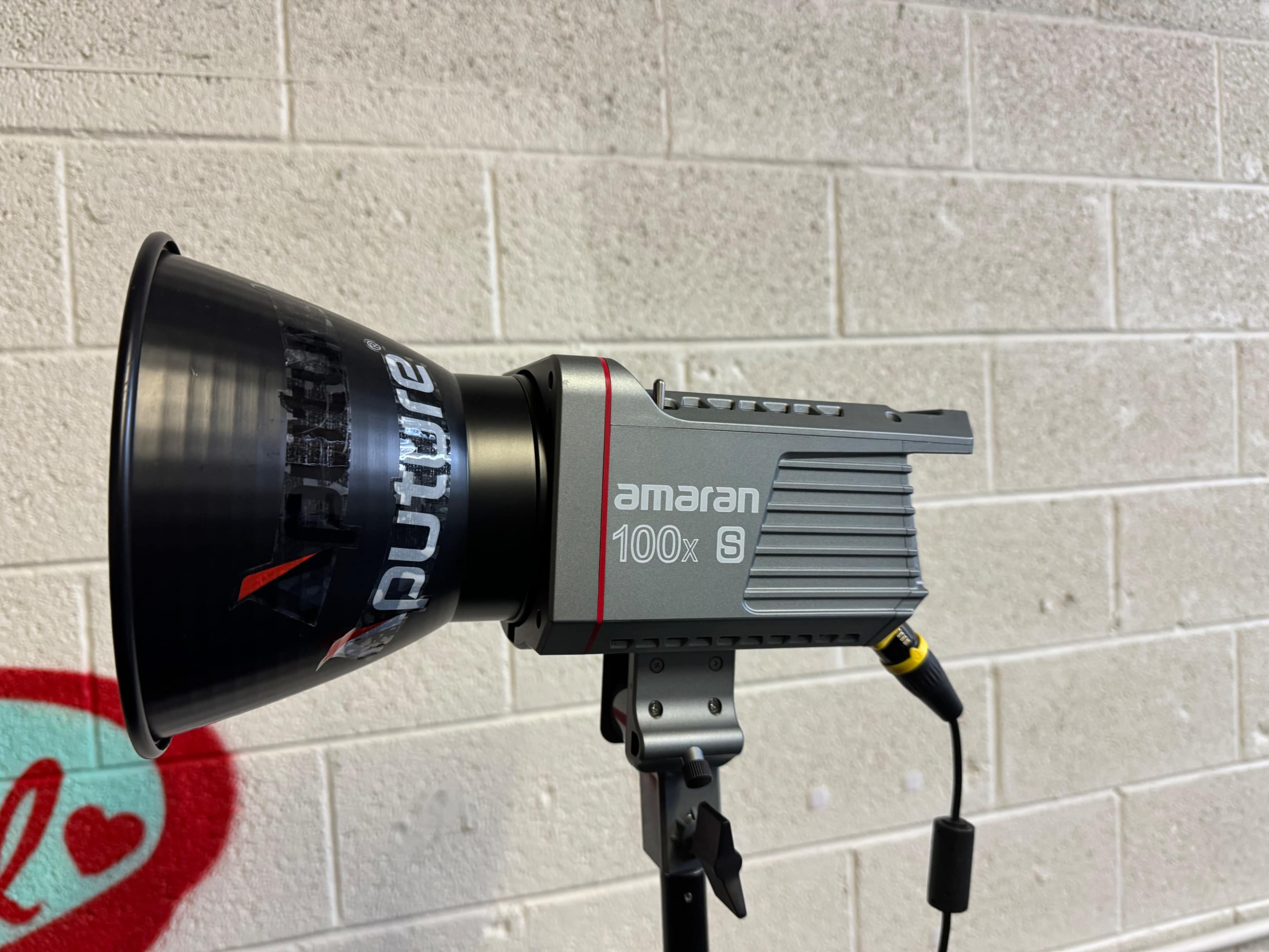 An amaran 100x S lighting fixture mounted on a stand against a concrete wall.