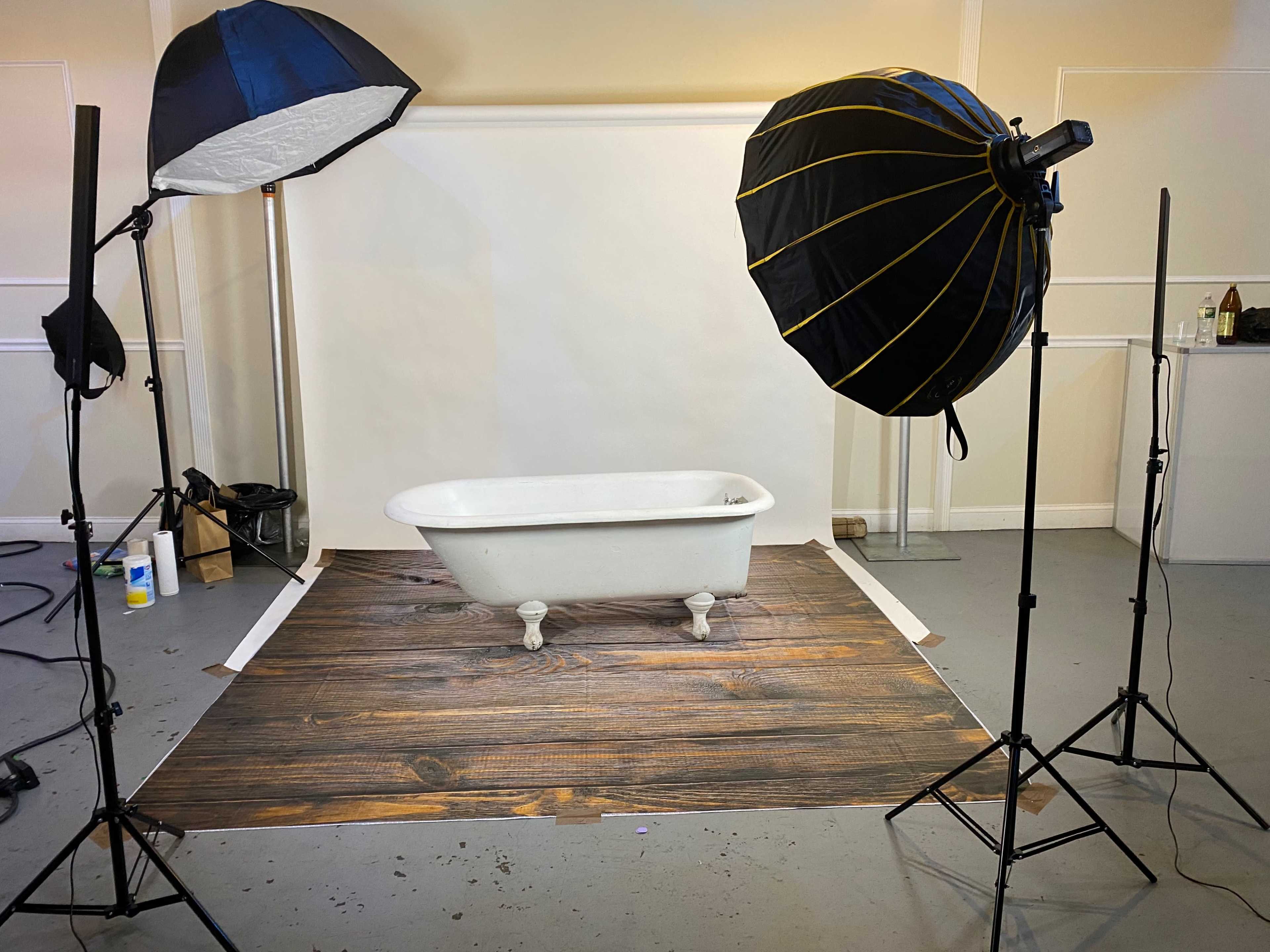 A white clawfoot bathtub positioned on a wooden floor backdrop, surrounded by two softbox lights in a studio setting.