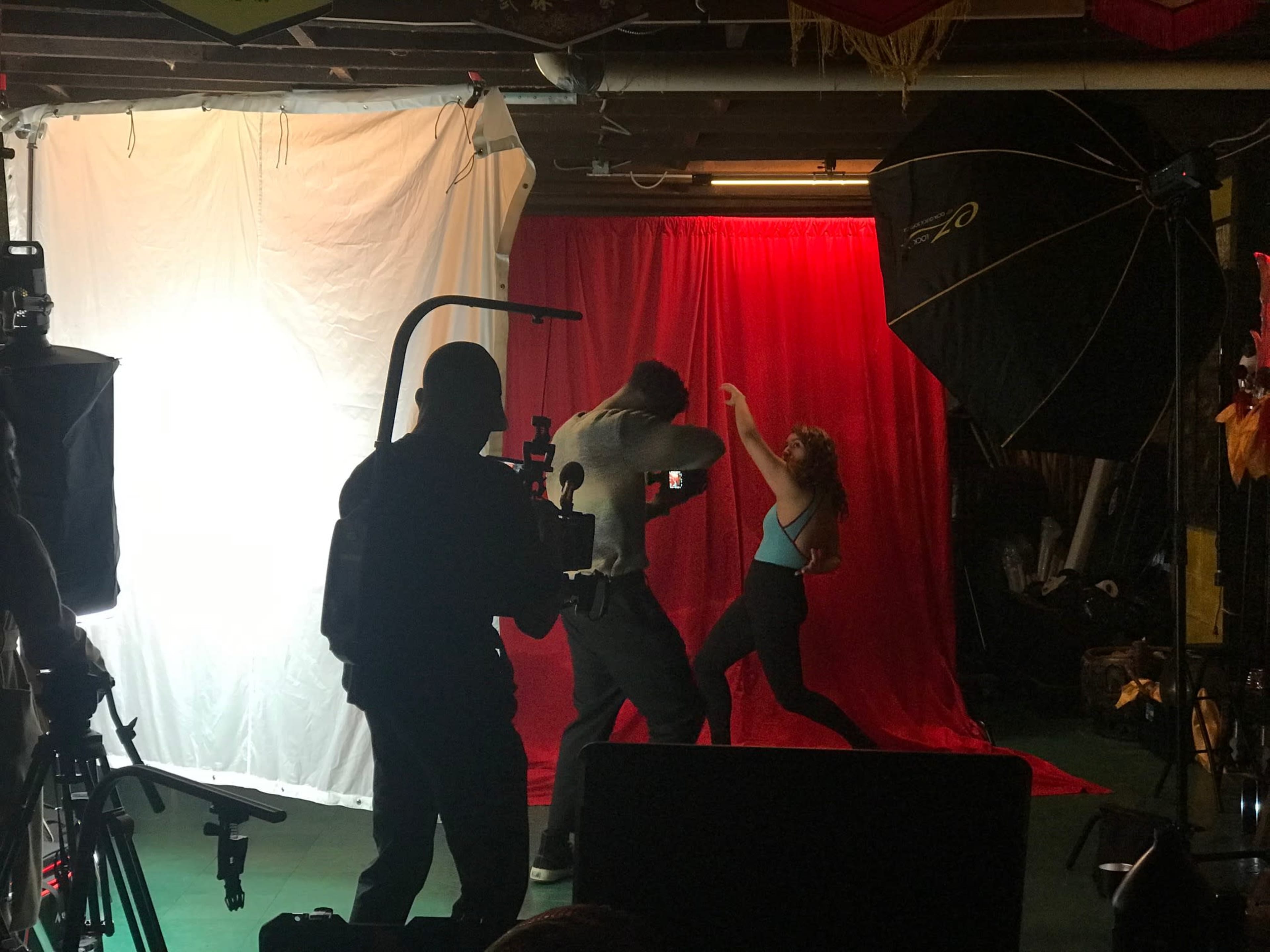 A photography shoot is in progress in a basement with a red backdrop, featuring a male model posing and a female model dancing, while a photographer captures the scene.