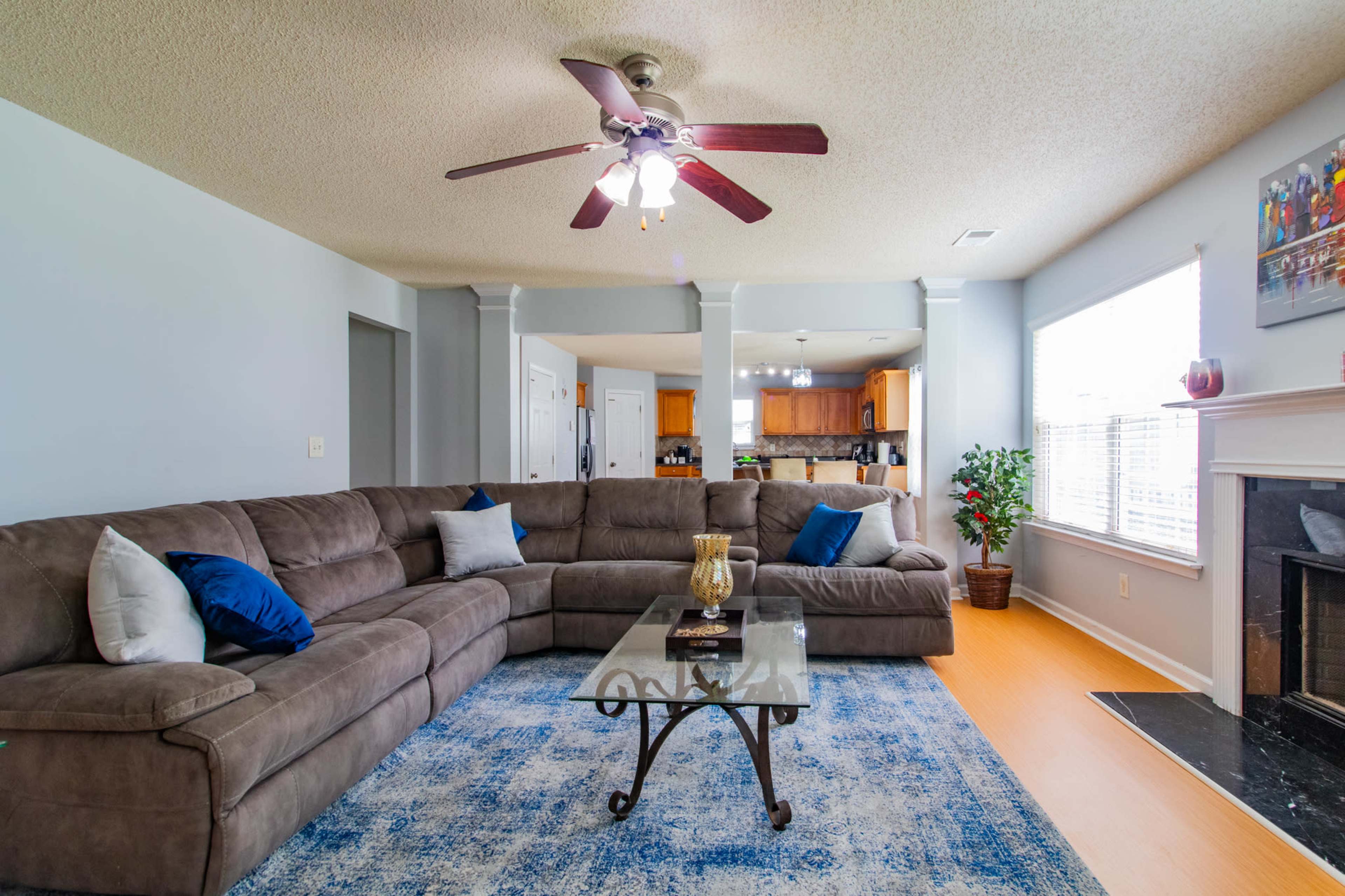 A spacious living room features a large sectional sofa, a glass coffee table, and a fireplace, with an open view into the kitchen area.