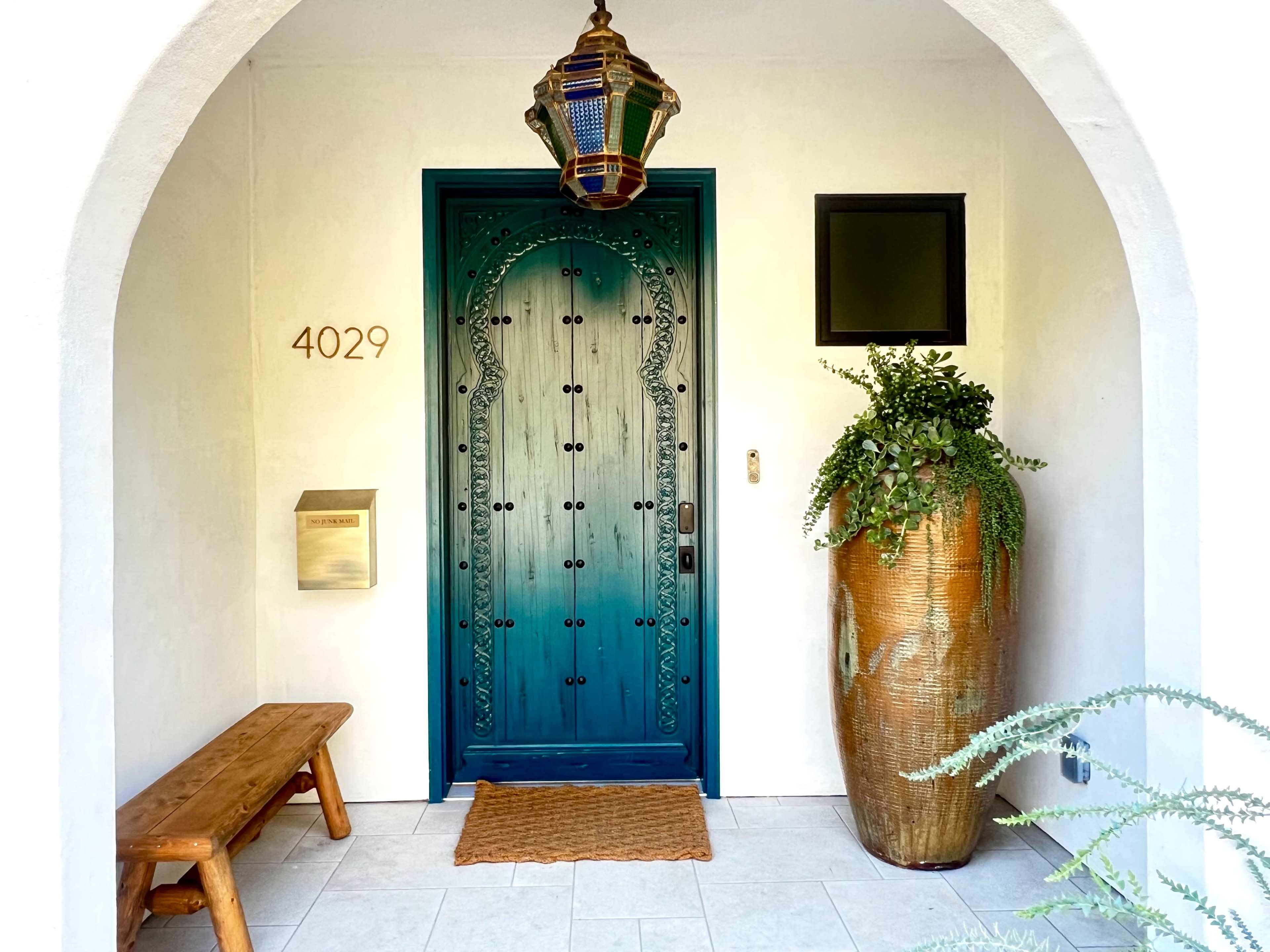 The entrance features a blue door with intricate designs, a hanging lantern, a large plant arrangement beside the door, and a wooden bench.