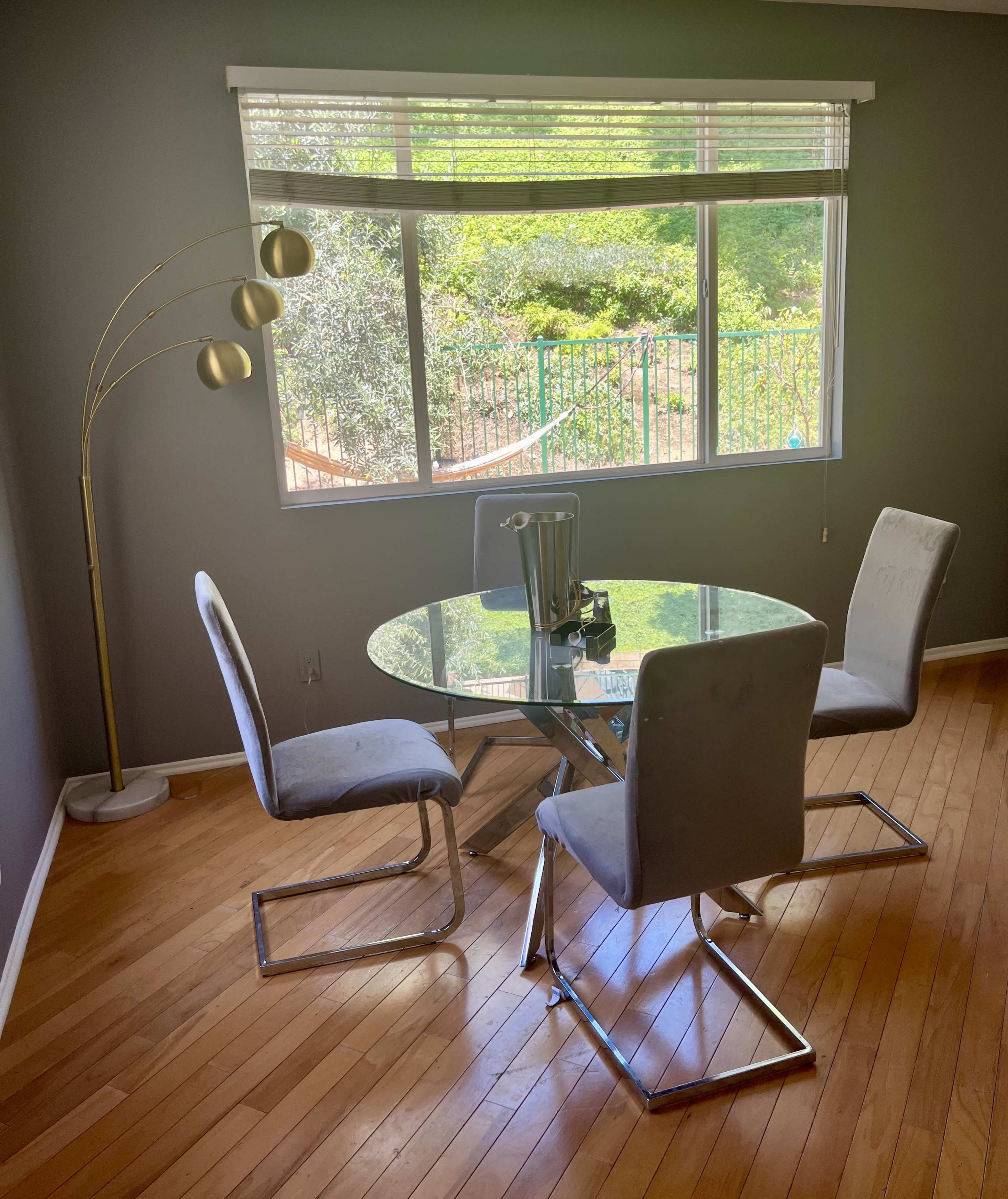 A circular glass dining table is set with three chairs in a well-lit room, featuring a large window that overlooks a garden.