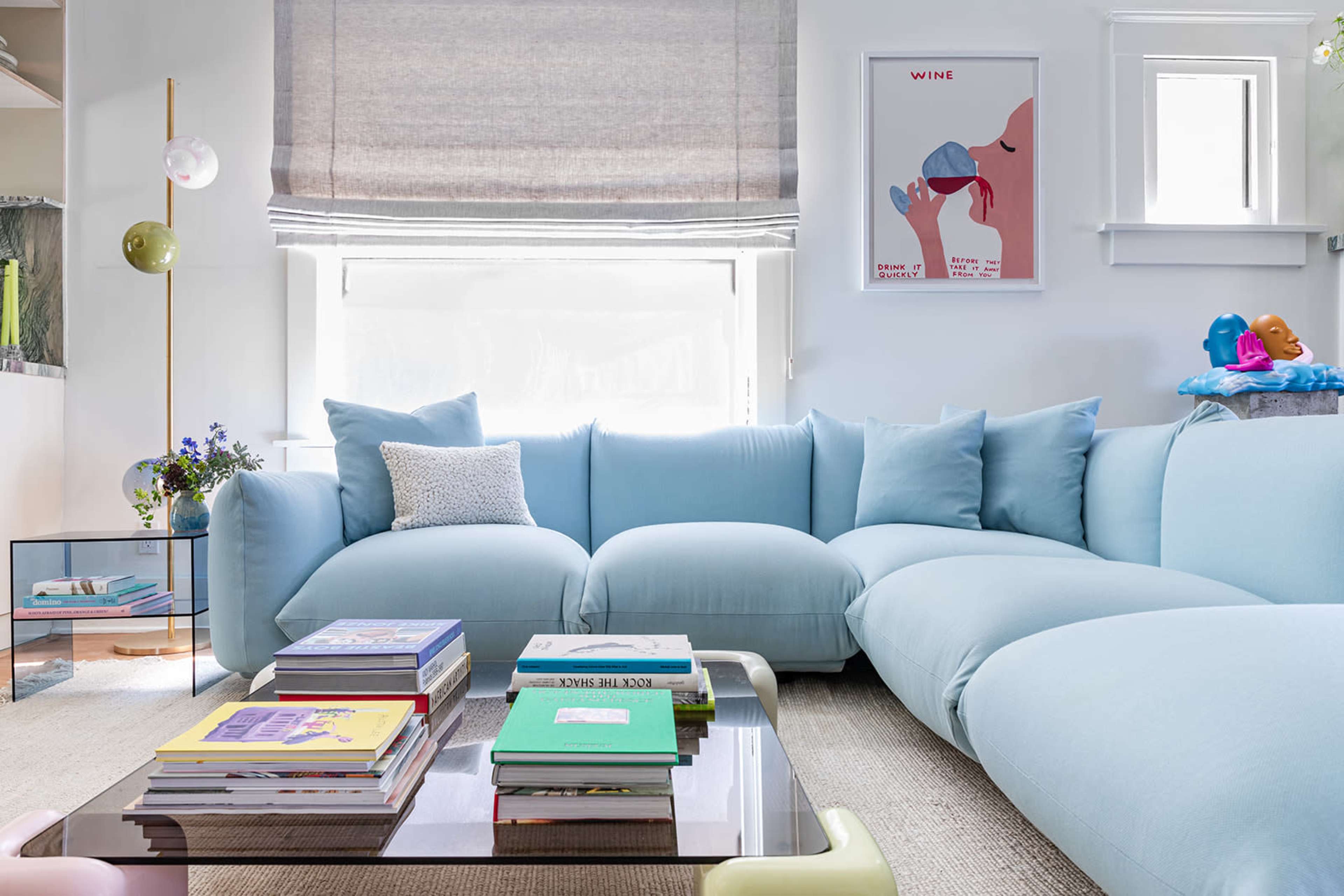 A light blue sectional sofa with a coffee table stacked with books, set against a backdrop of a window with shades and a framed poster on the wall.