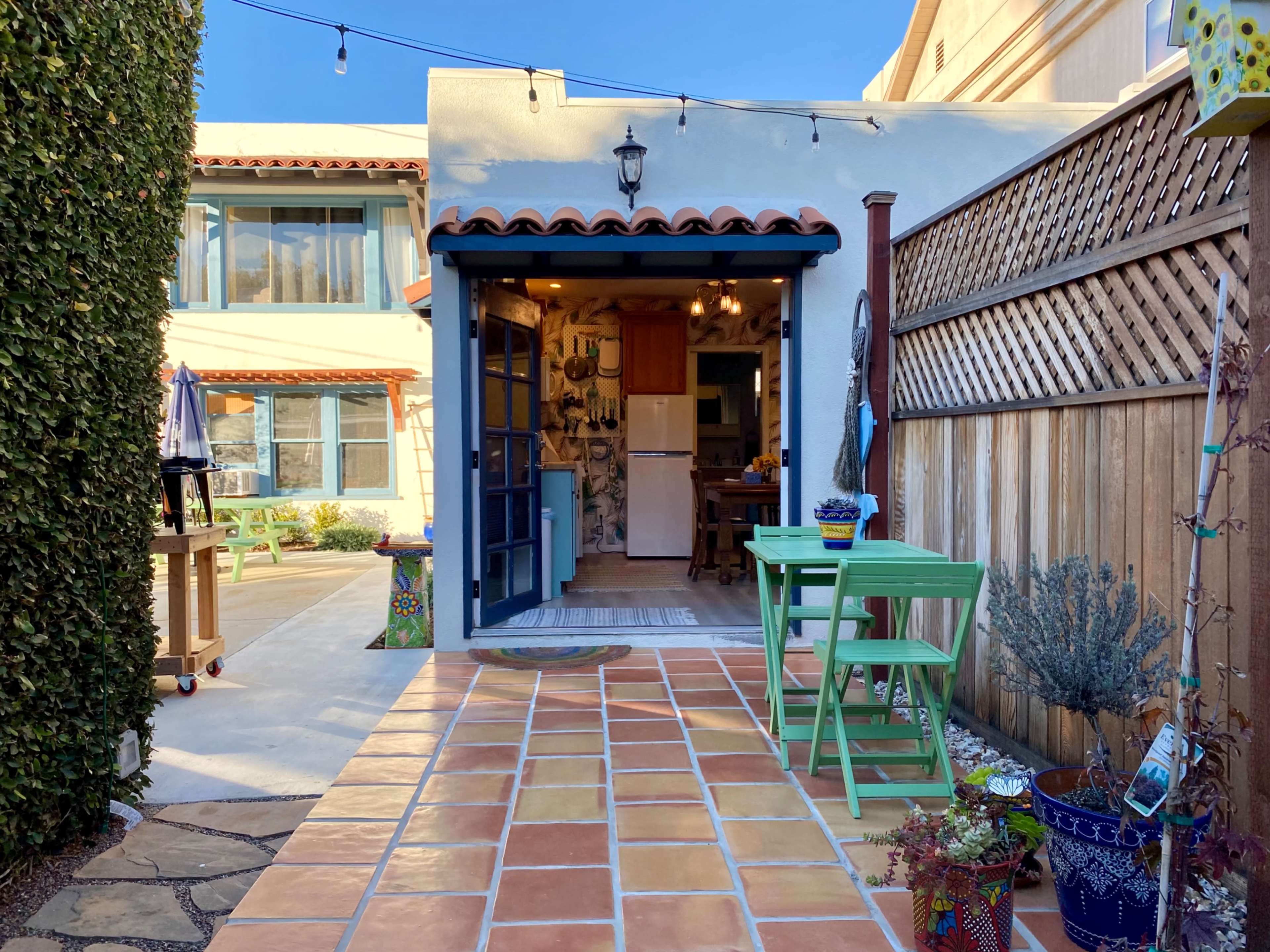 The image shows a quaint outdoor patio with a tiled floor, green furniture, and an open doorway leading into a kitchen area surrounded by greenery and a wooden fence.