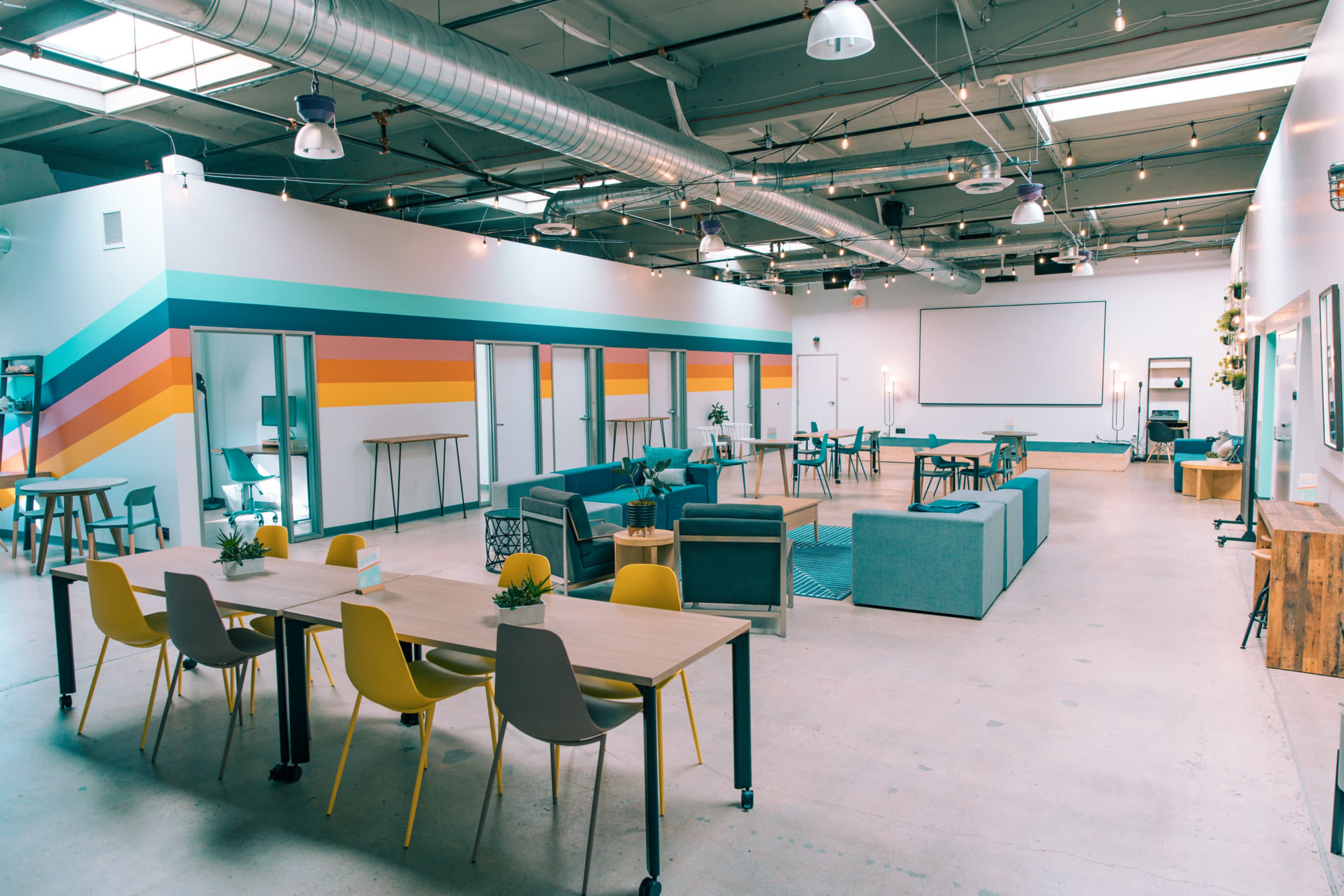 The image shows a spacious, modern meeting area with colorful accent walls, various seating arrangements, and multiple tables for collaboration.