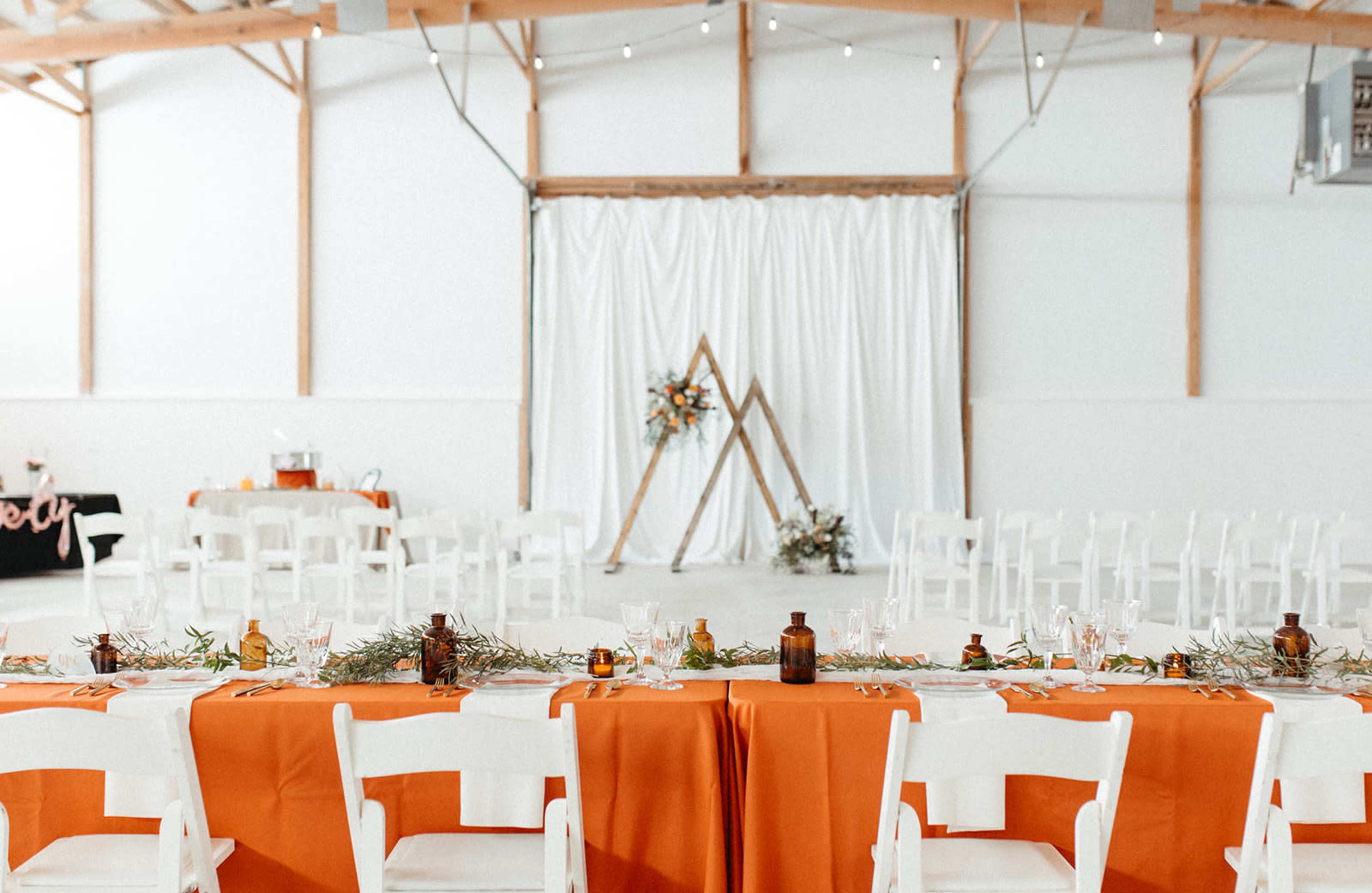 A long table with orange linens is set up in a spacious event venue, featuring a backdrop of white drapes and a wooden frame adorned with greenery and flowers.