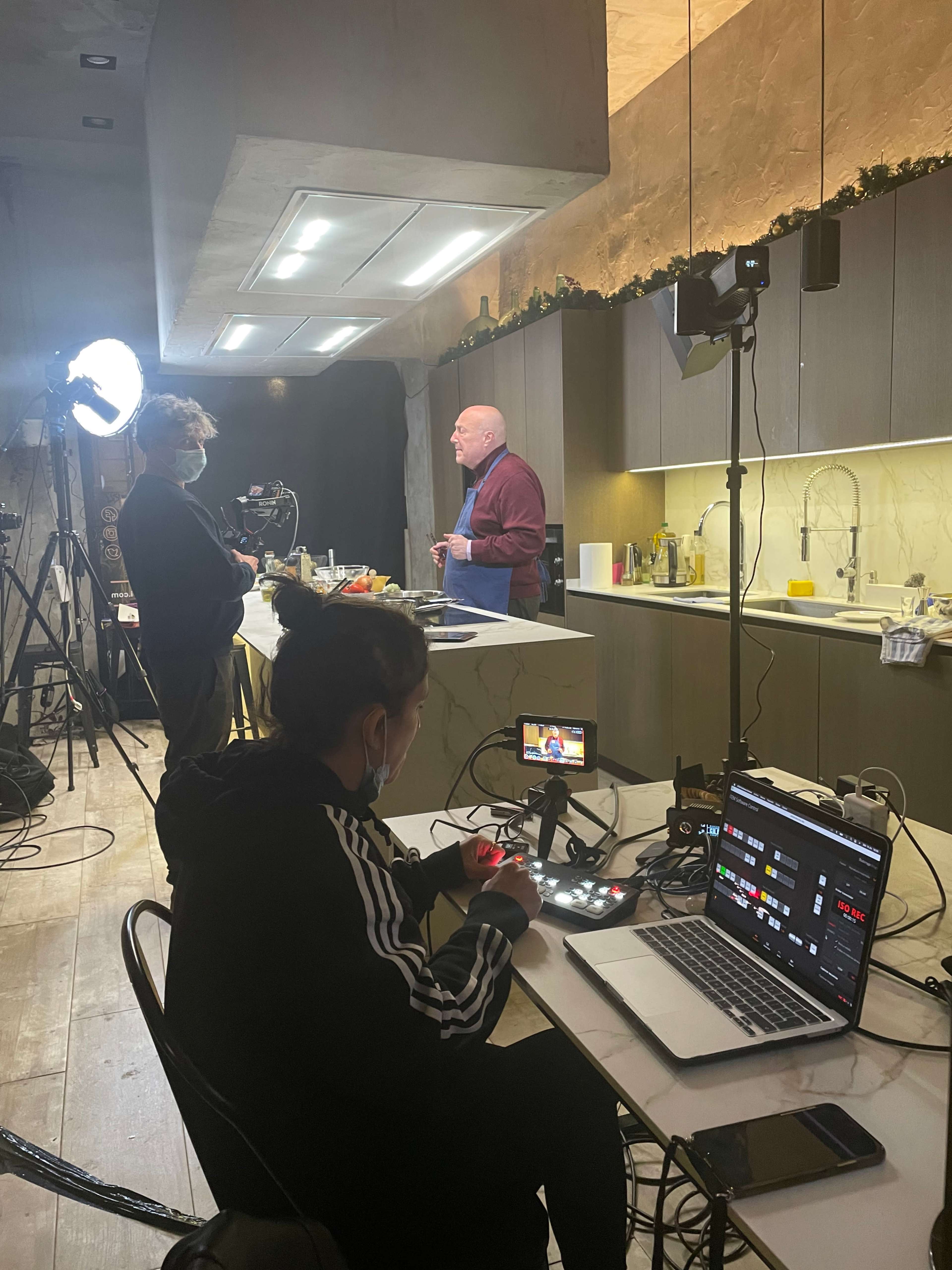 A man stands at a kitchen counter while another person operates a camera and equipment in a well-lit studio space.