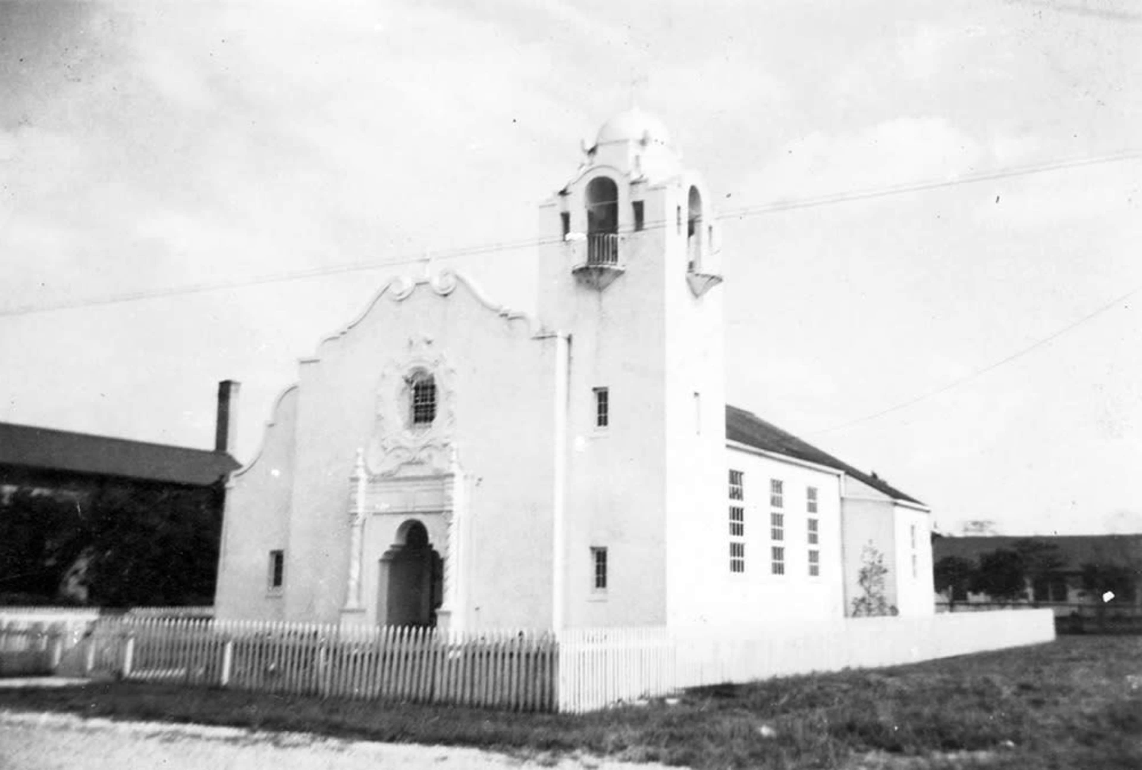 A white, single-story building with a decorative facade and a small tower, surrounded by a white picket fence.