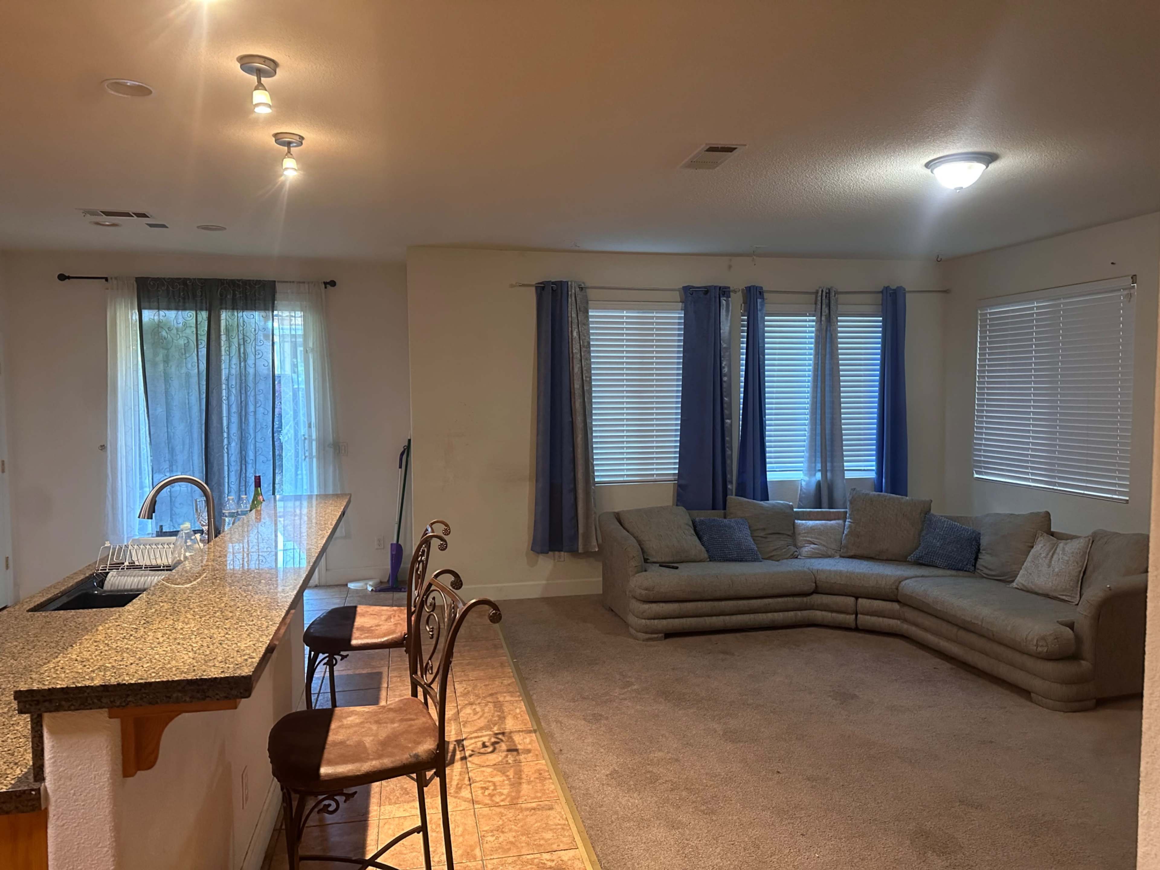 A living room with a grey sectional sofa, a bar area with two stools, and large windows covered with curtains.