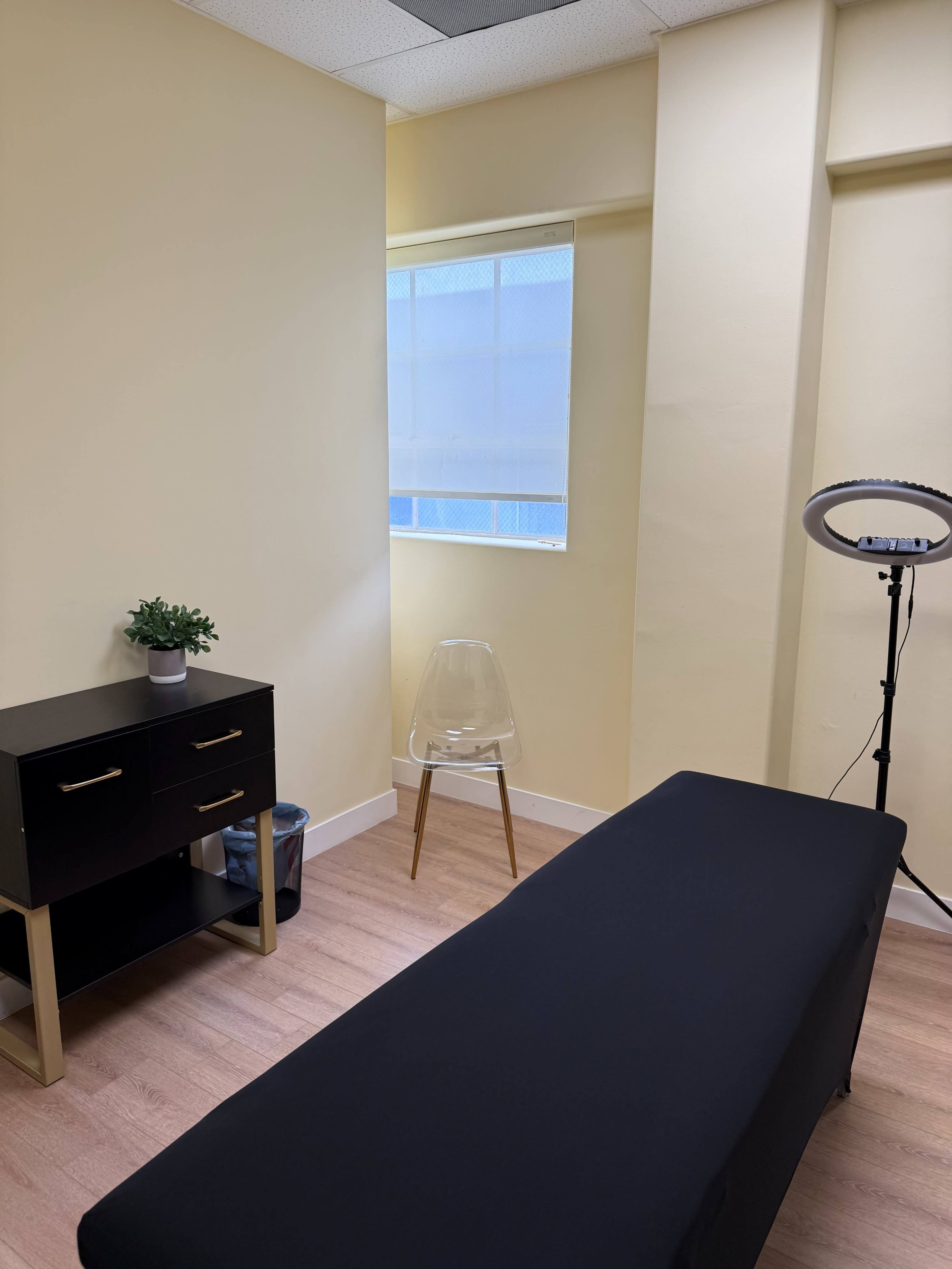 The image shows an interior room with a window, a black massage table, a clear chair, a small black cabinet, and a ring light.