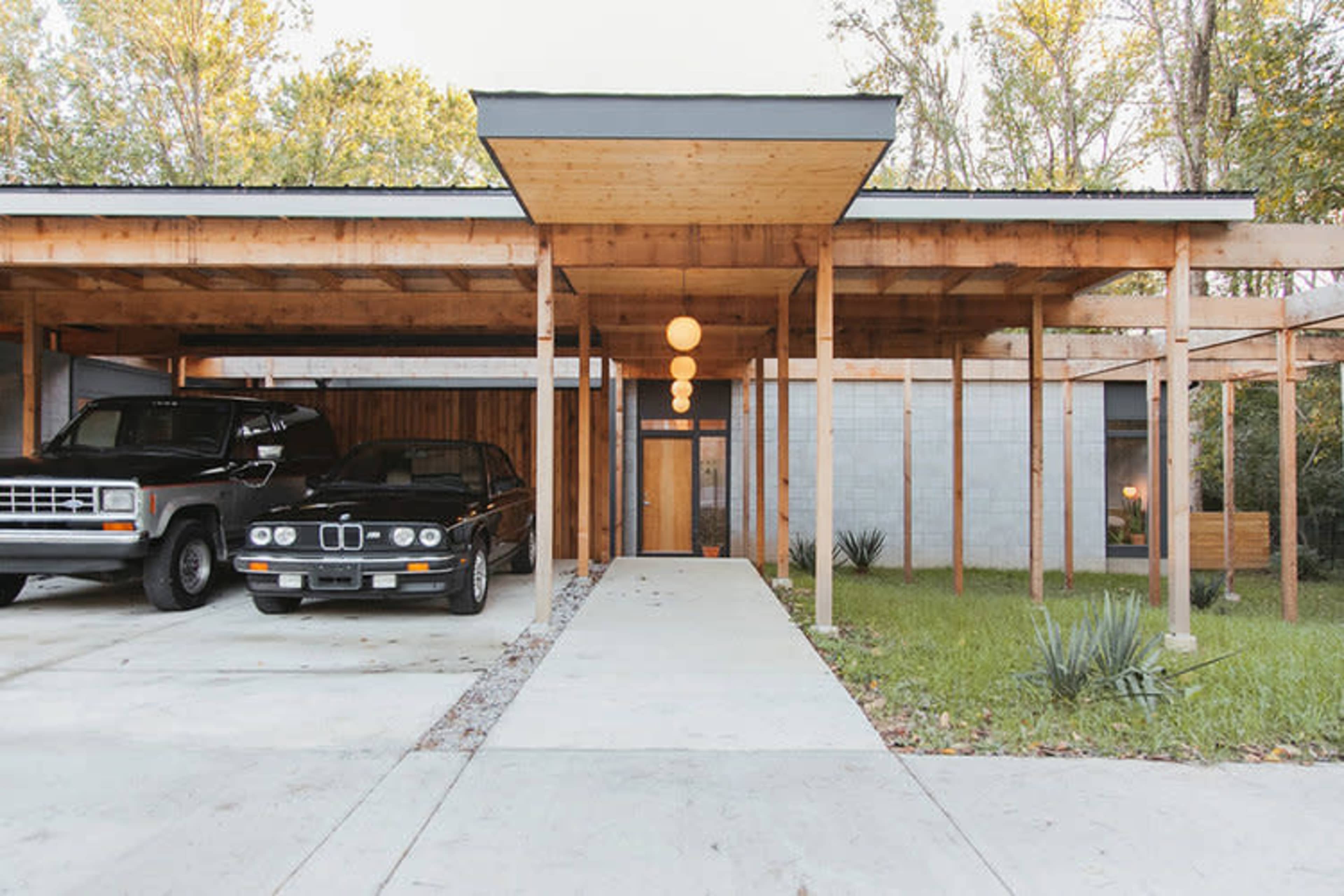 A modern house with a spacious carport, featuring two parked vehicles on a concrete driveway and surrounded by greenery.