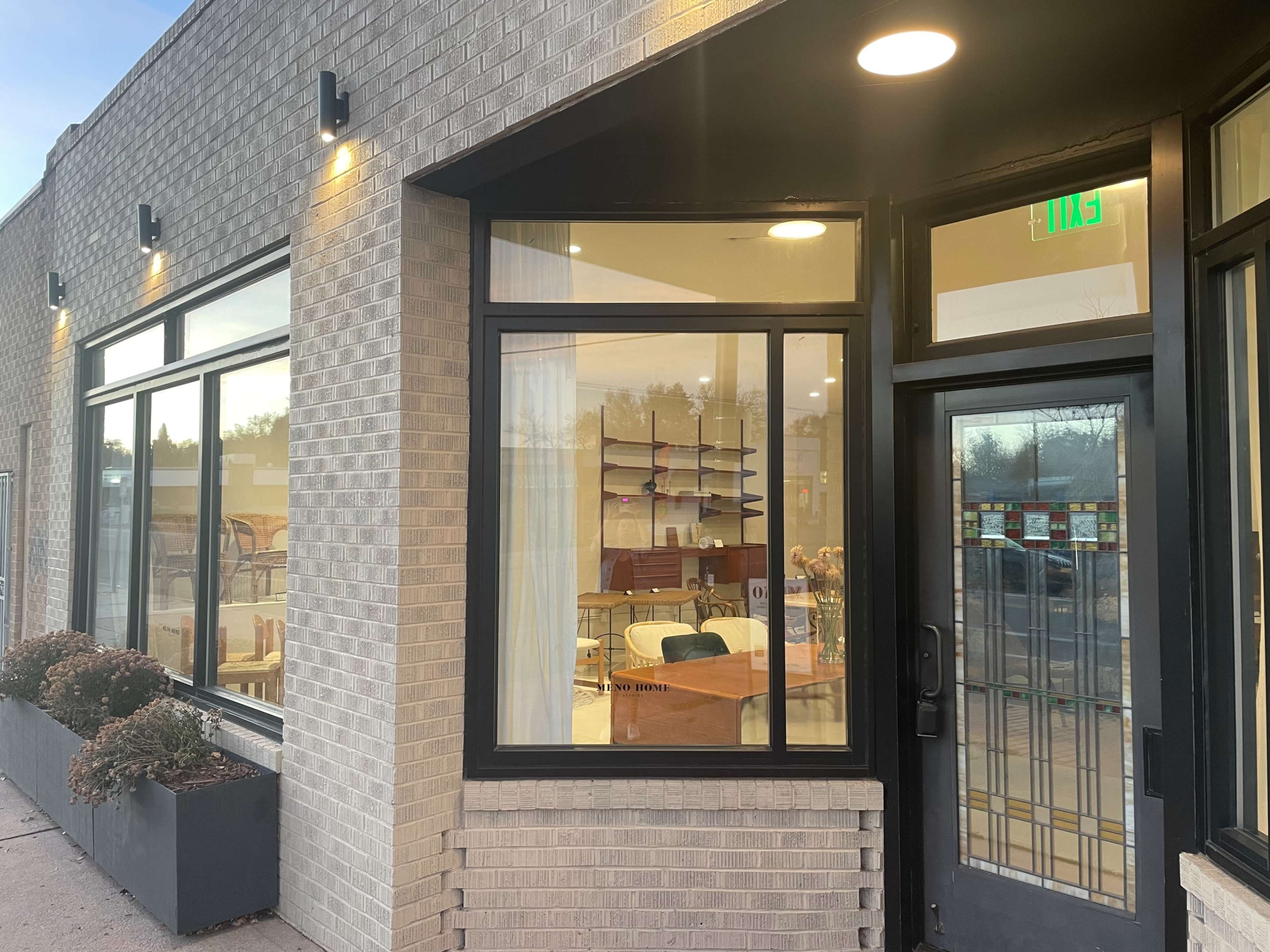 A modern building exterior with large windows, showcasing interior furniture and decor, and a glass door with a visible exit sign.