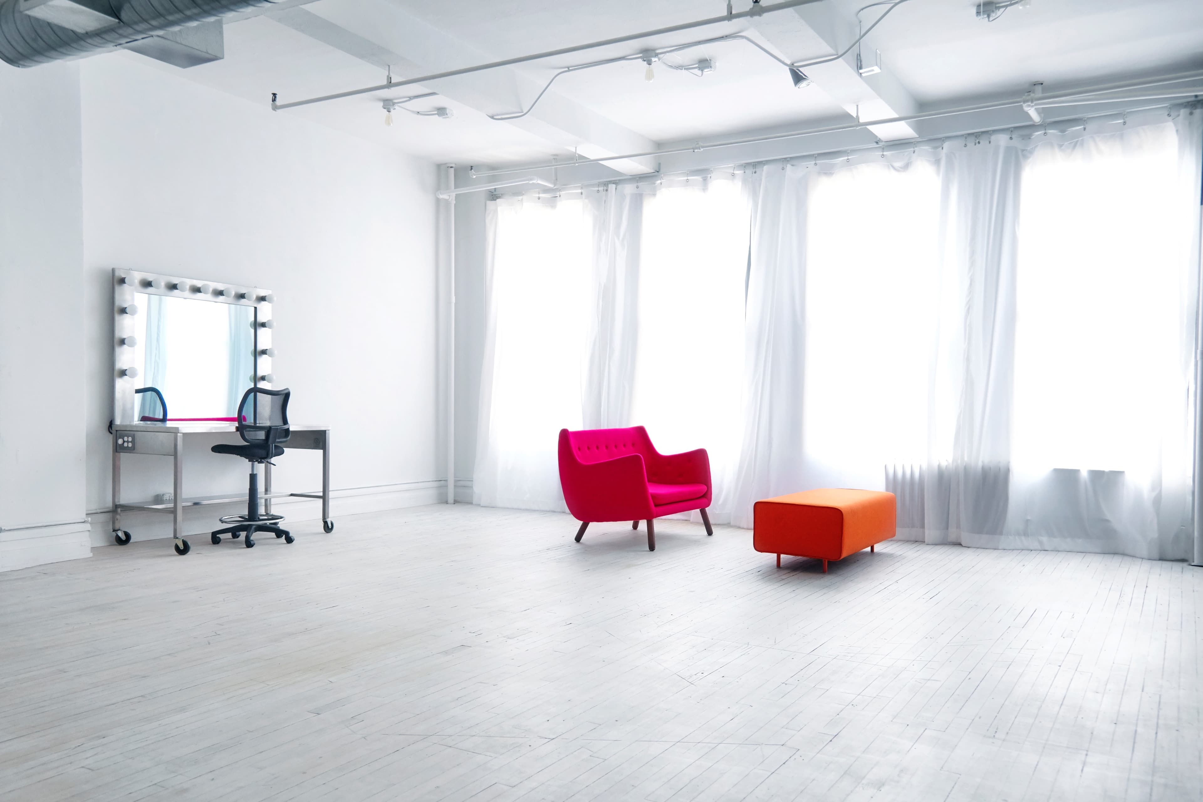 A bright, minimalist studio features a pink armchair, an orange ottoman, and a mirrored vanity with a rolling chair, all complemented by large windows draped with white curtains.