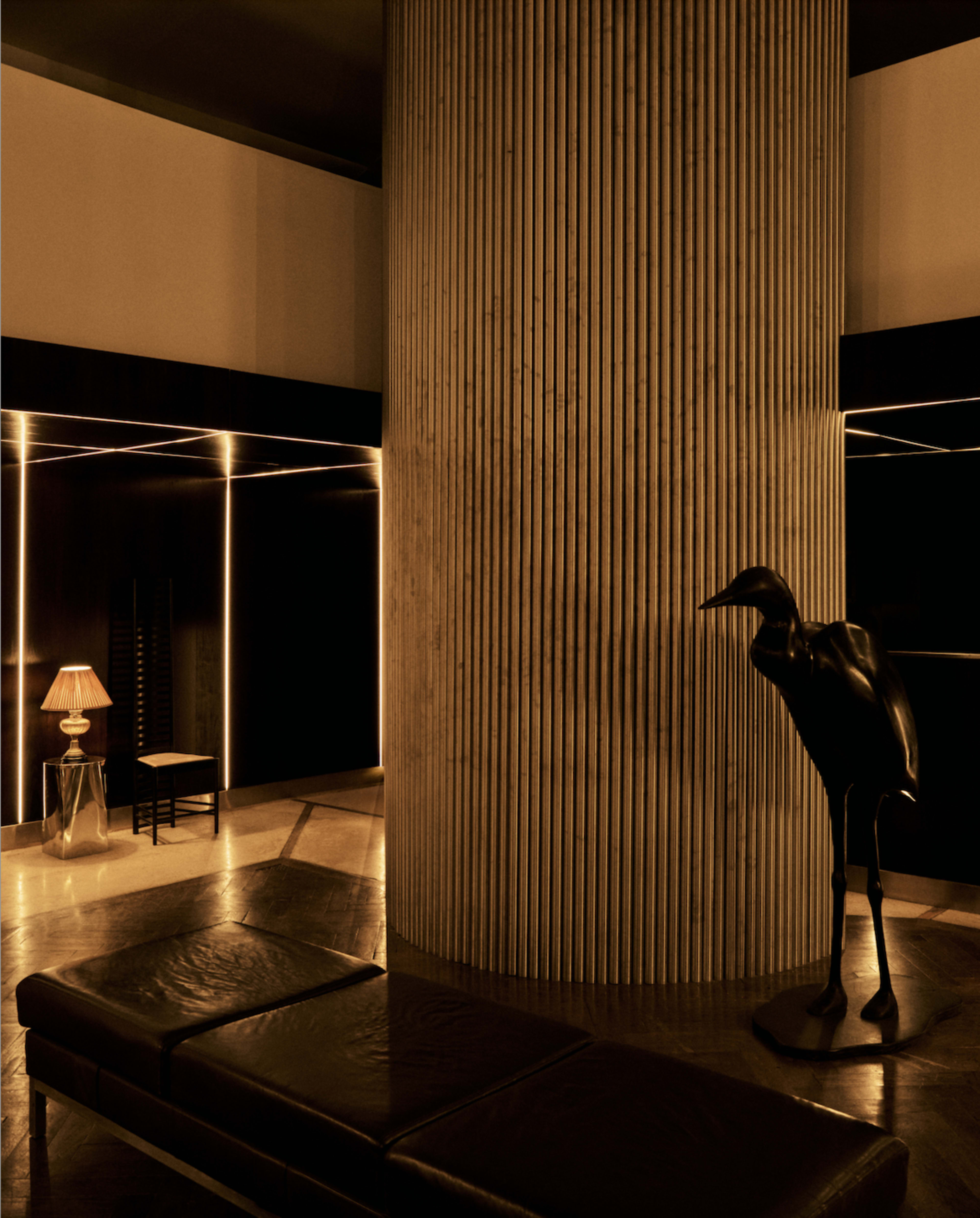 The image shows a minimalistic interior space featuring a tall, textured column, a dark sculpture resembling a bird, and a chair illuminated by soft lighting.