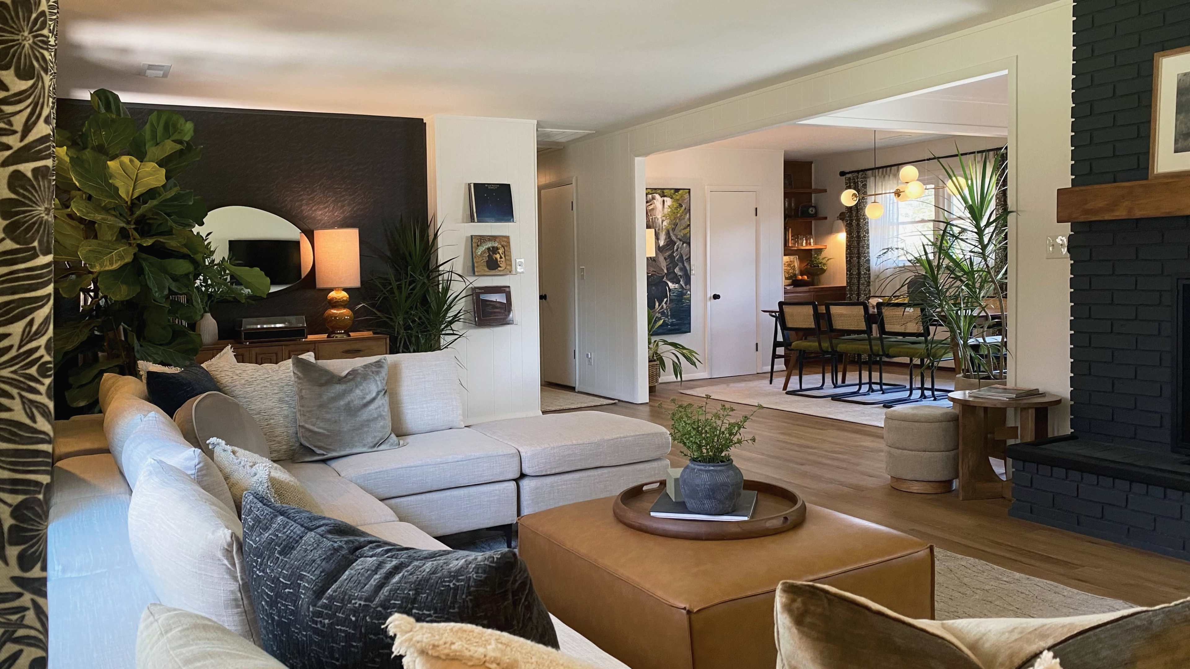 The image shows a modern living room with a sectional sofa, a round mirror, and plants, leading into a dining area with a table and chairs.