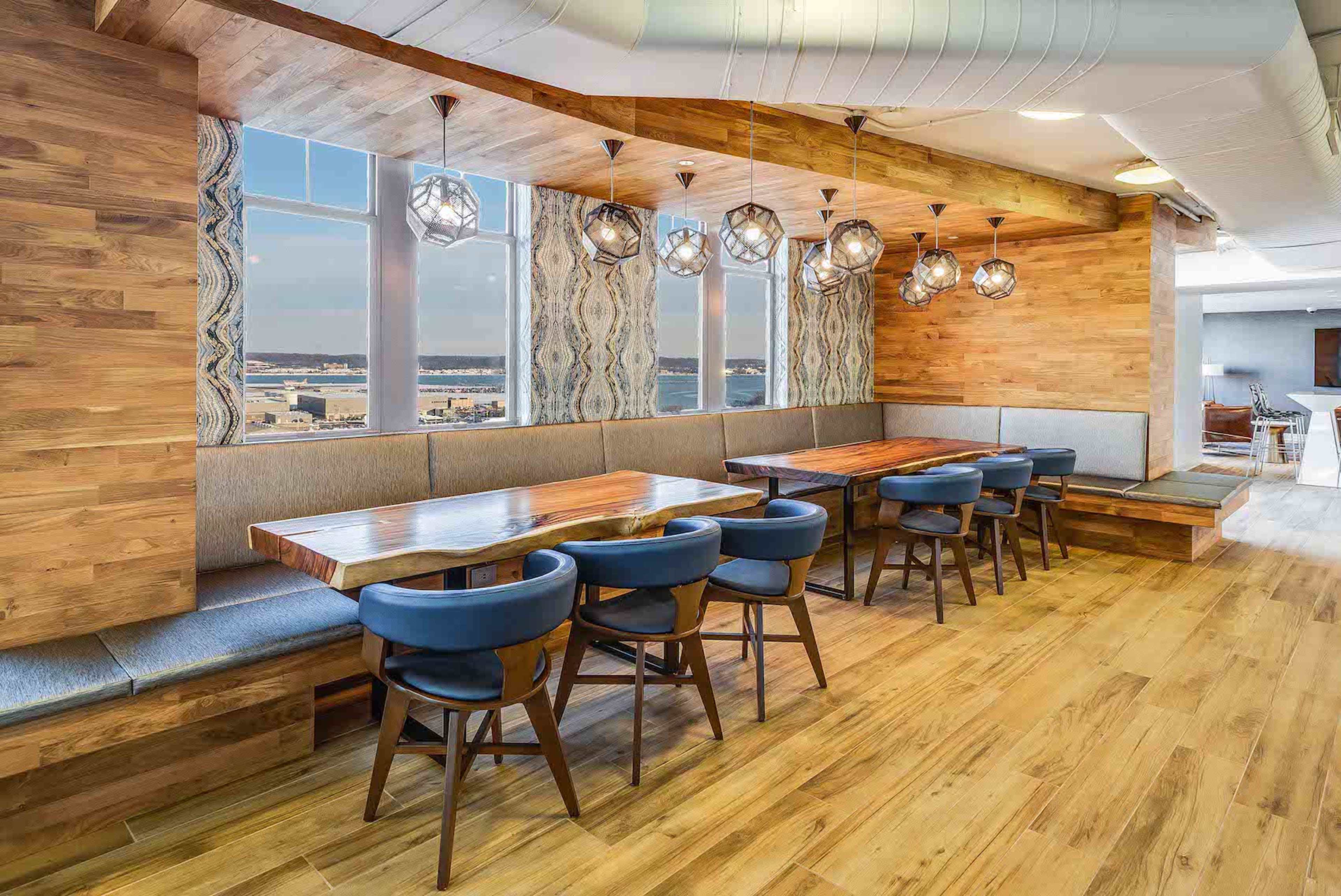 A modern dining area with wooden walls, large windows, and a row of tables surrounded by blue chairs.