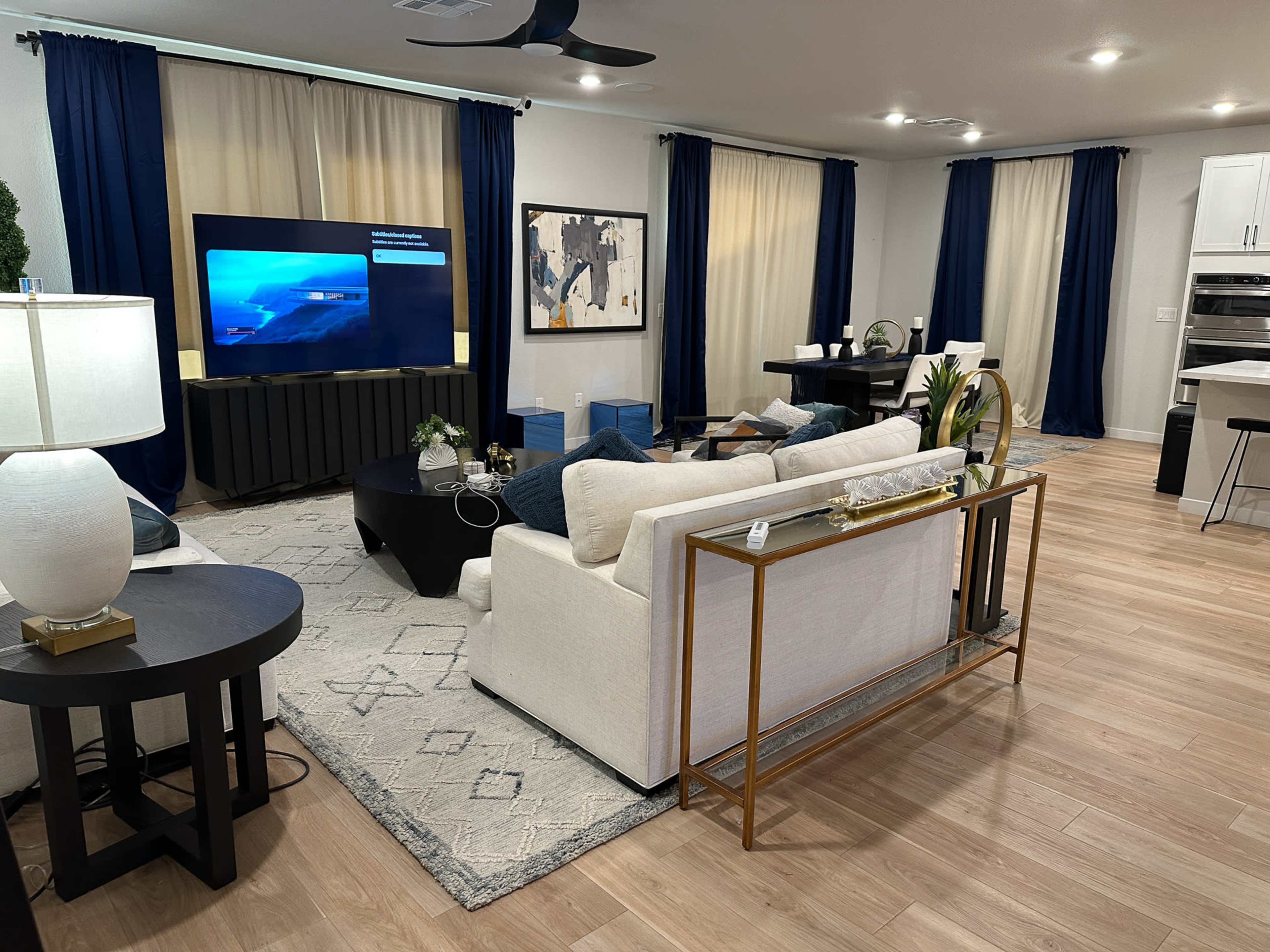 The image shows a modern living room featuring a light-colored sofa, a black coffee table, a large television, and blue curtains.