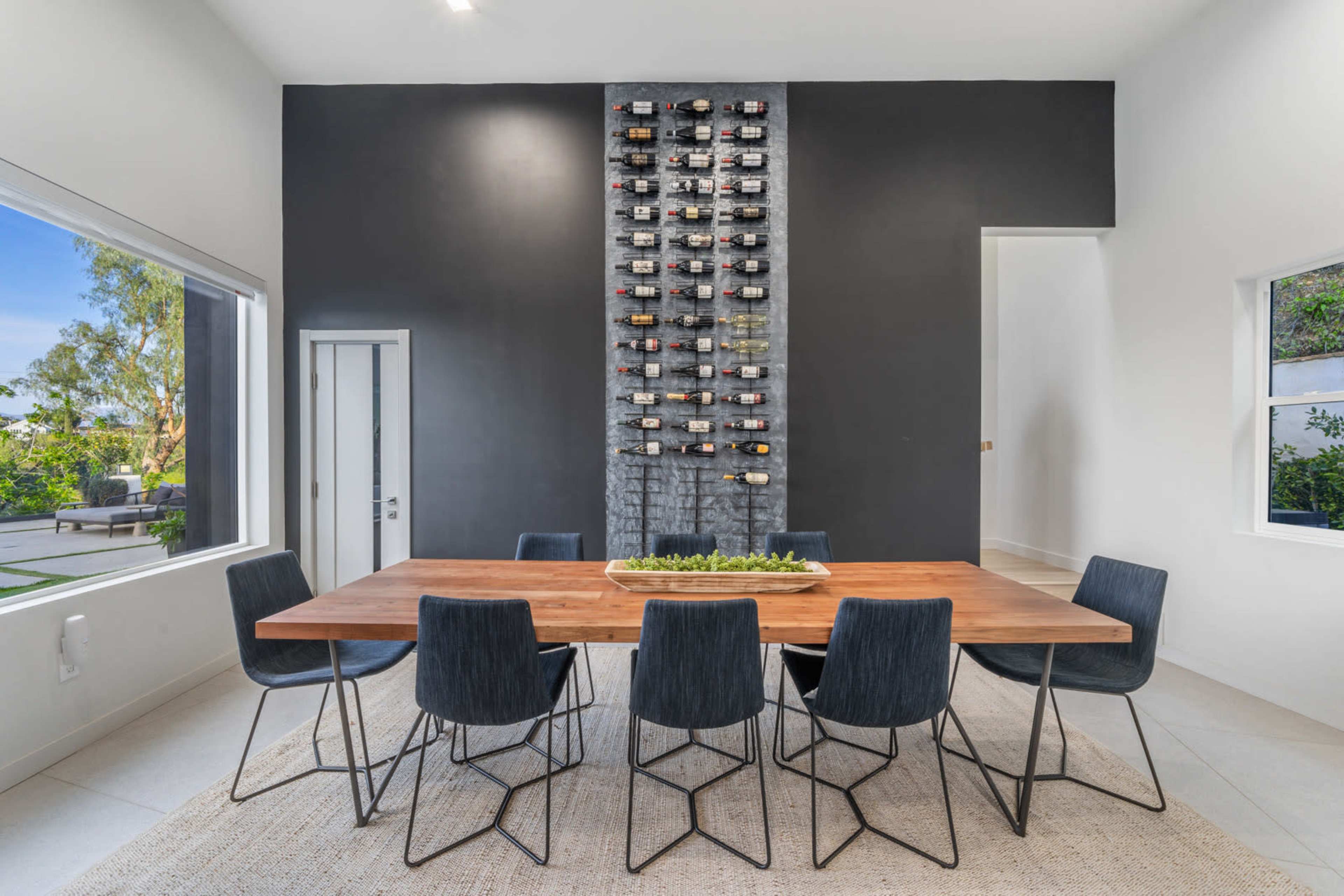A wooden dining table surrounded by six black chairs is set against a wall adorned with a wine rack in a modern interior space.