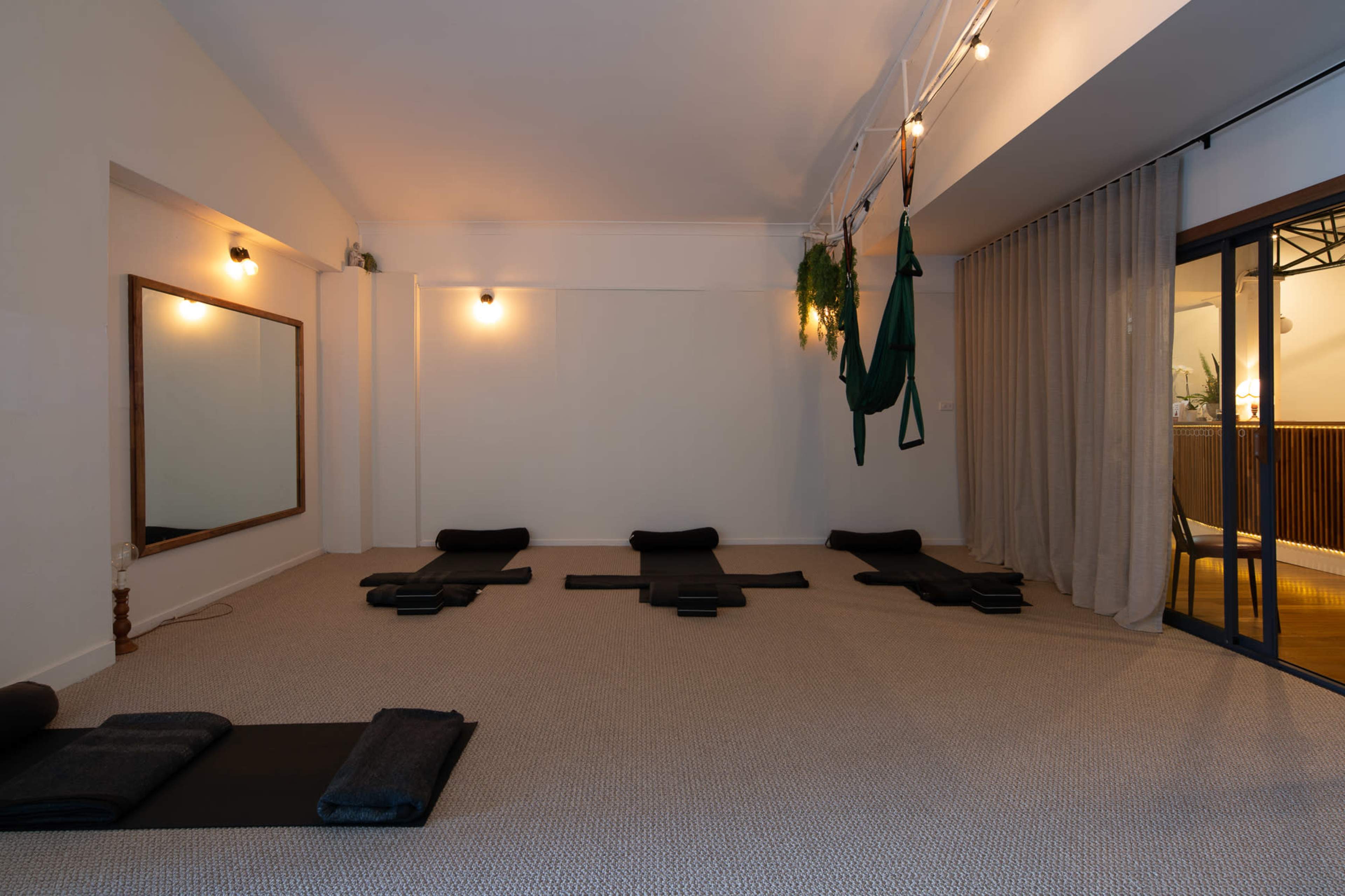 A spacious yoga studio with several black mats arranged on a carpeted floor, a large mirror on one wall, and curtains partially separating the space.