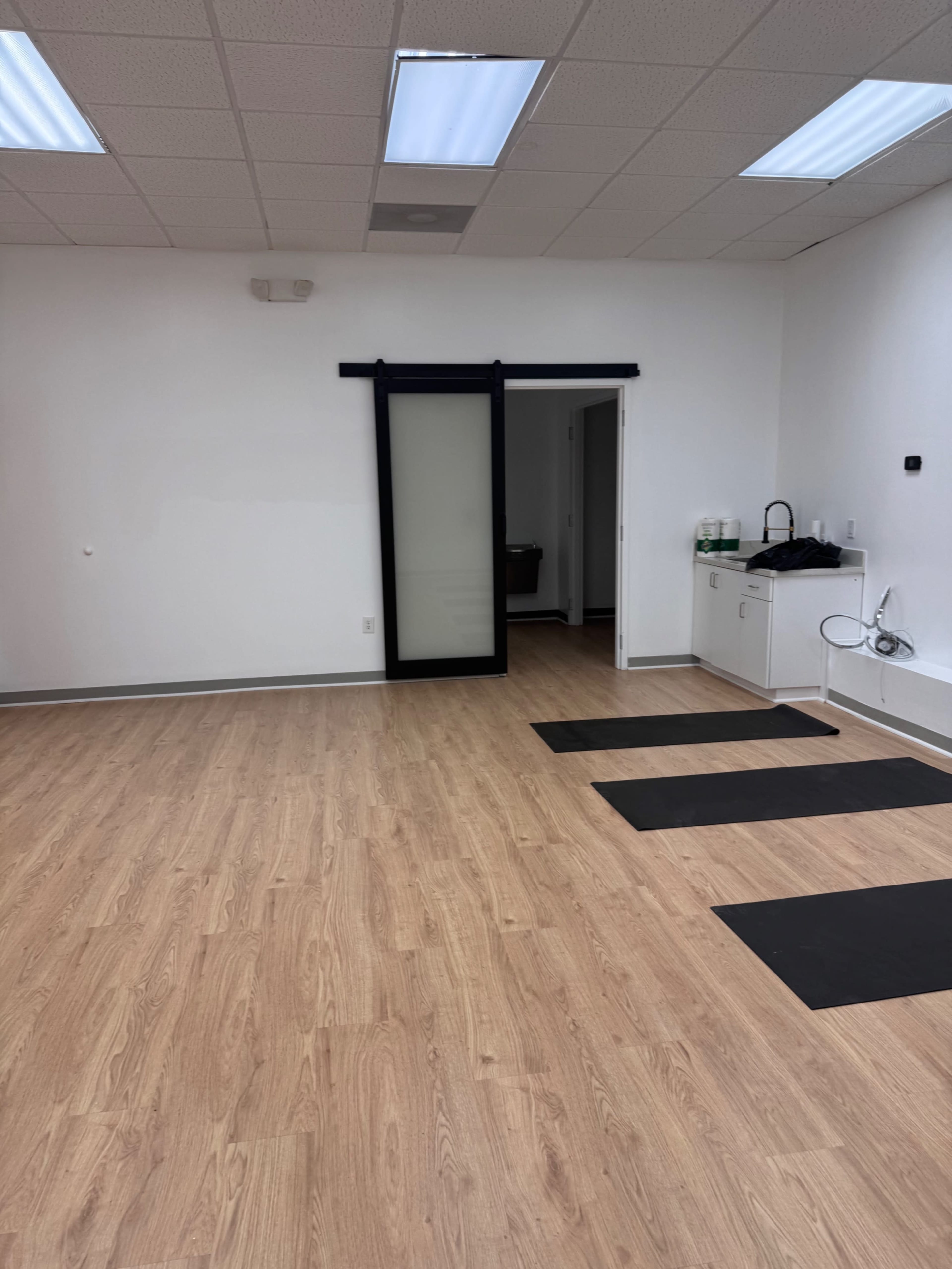 The image shows an empty room with wooden flooring, a sliding black door on the left, and three black exercise mats laid out on the floor.