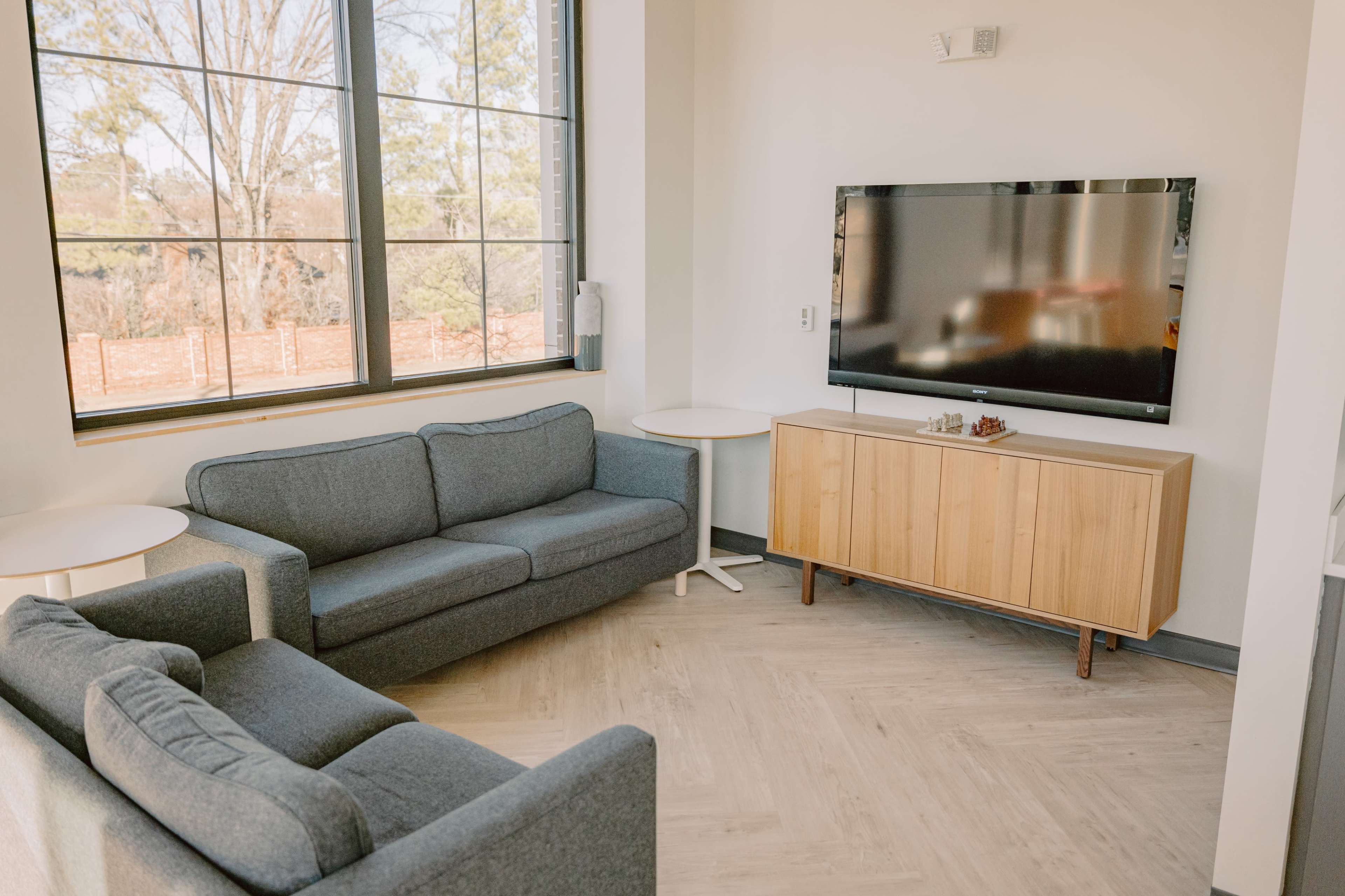 The living area features two grey sofas facing a wooden TV stand with a television mounted on the wall, alongside a large window providing natural light.