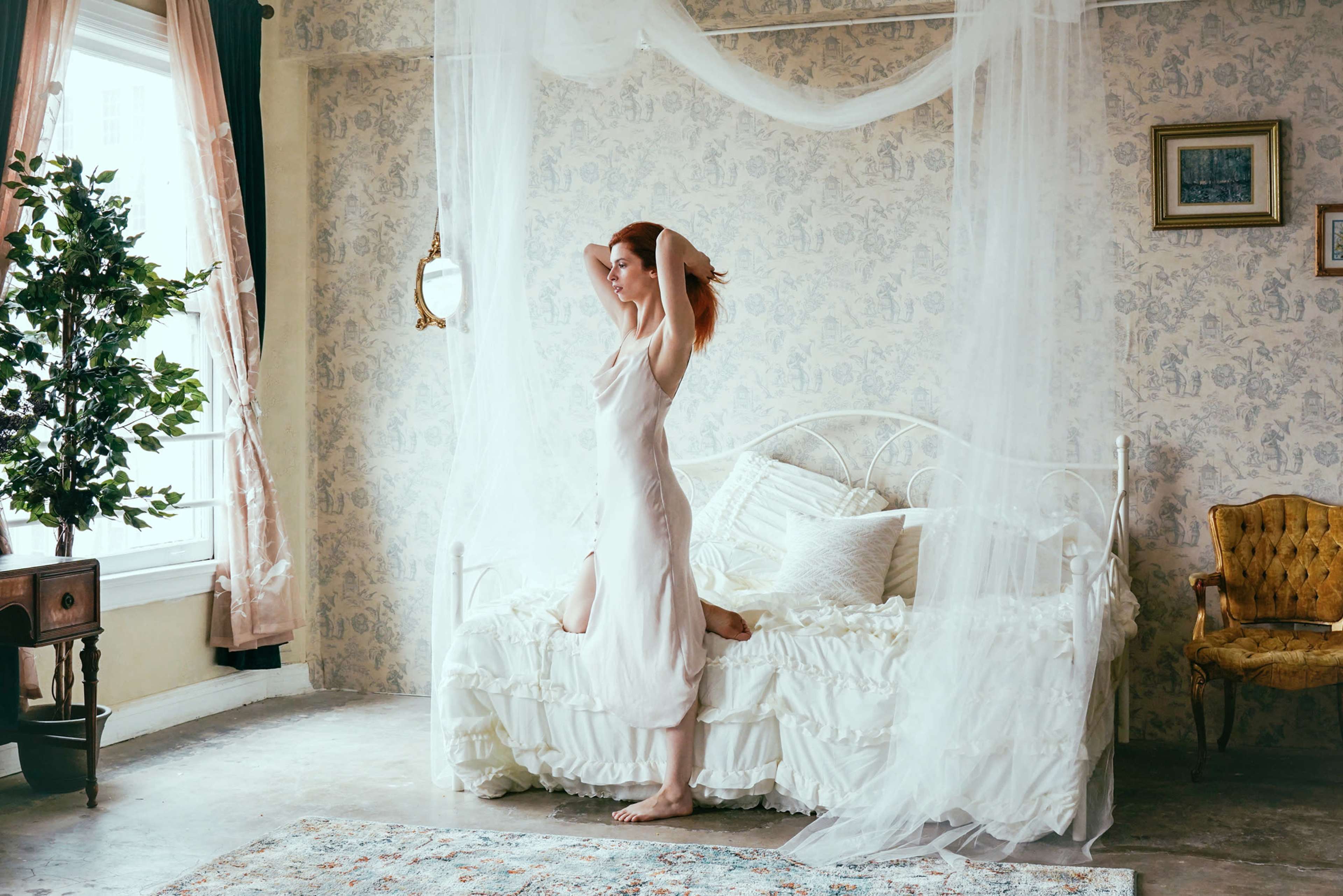 A woman stands on a bed in a softly lit room, surrounded by sheer curtains and vintage decor.