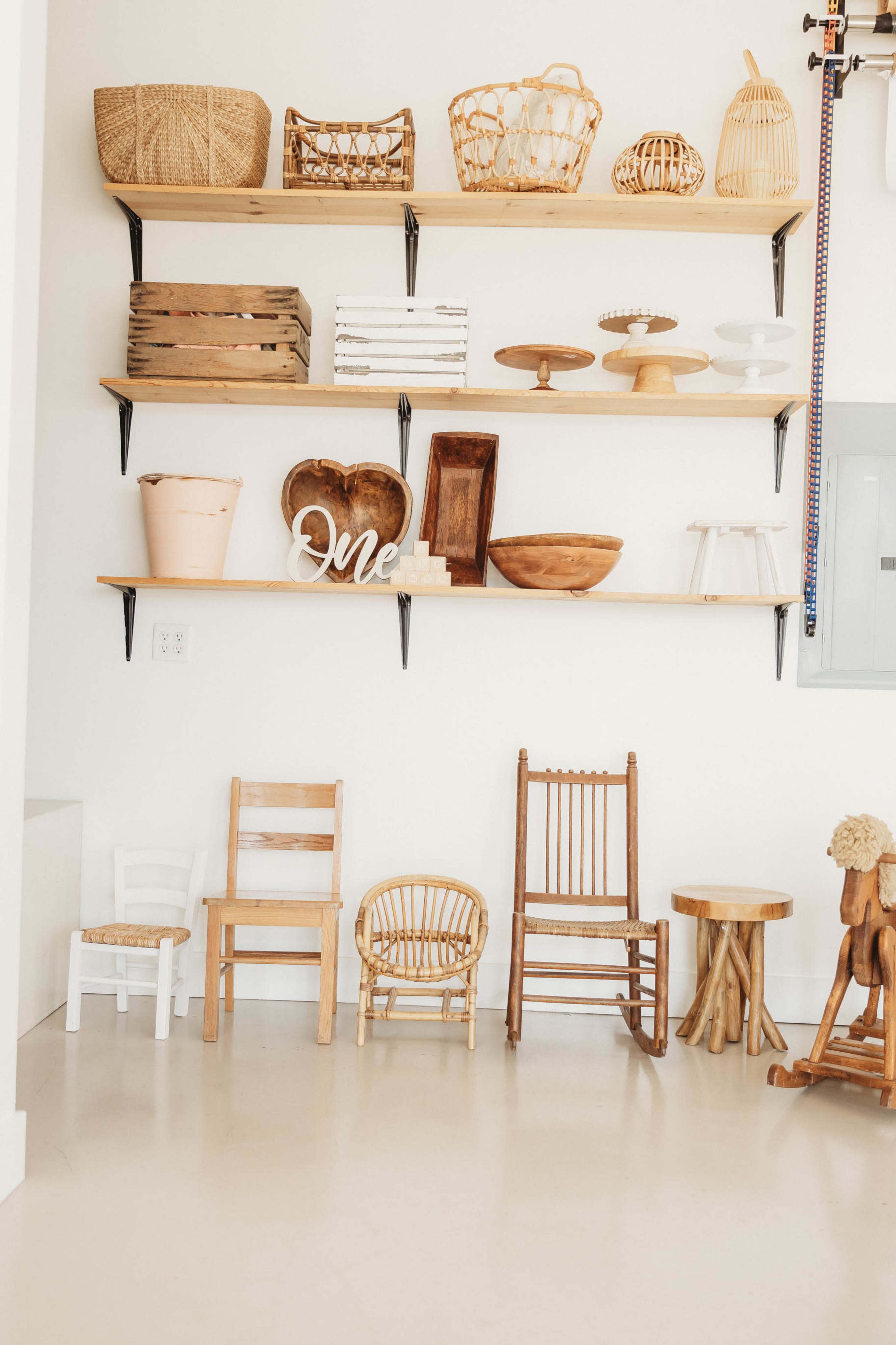 The image shows a wall with wooden shelves displaying various woven baskets and wooden bowls above a collection of small furniture pieces, including chairs and stools, arranged on the floor.