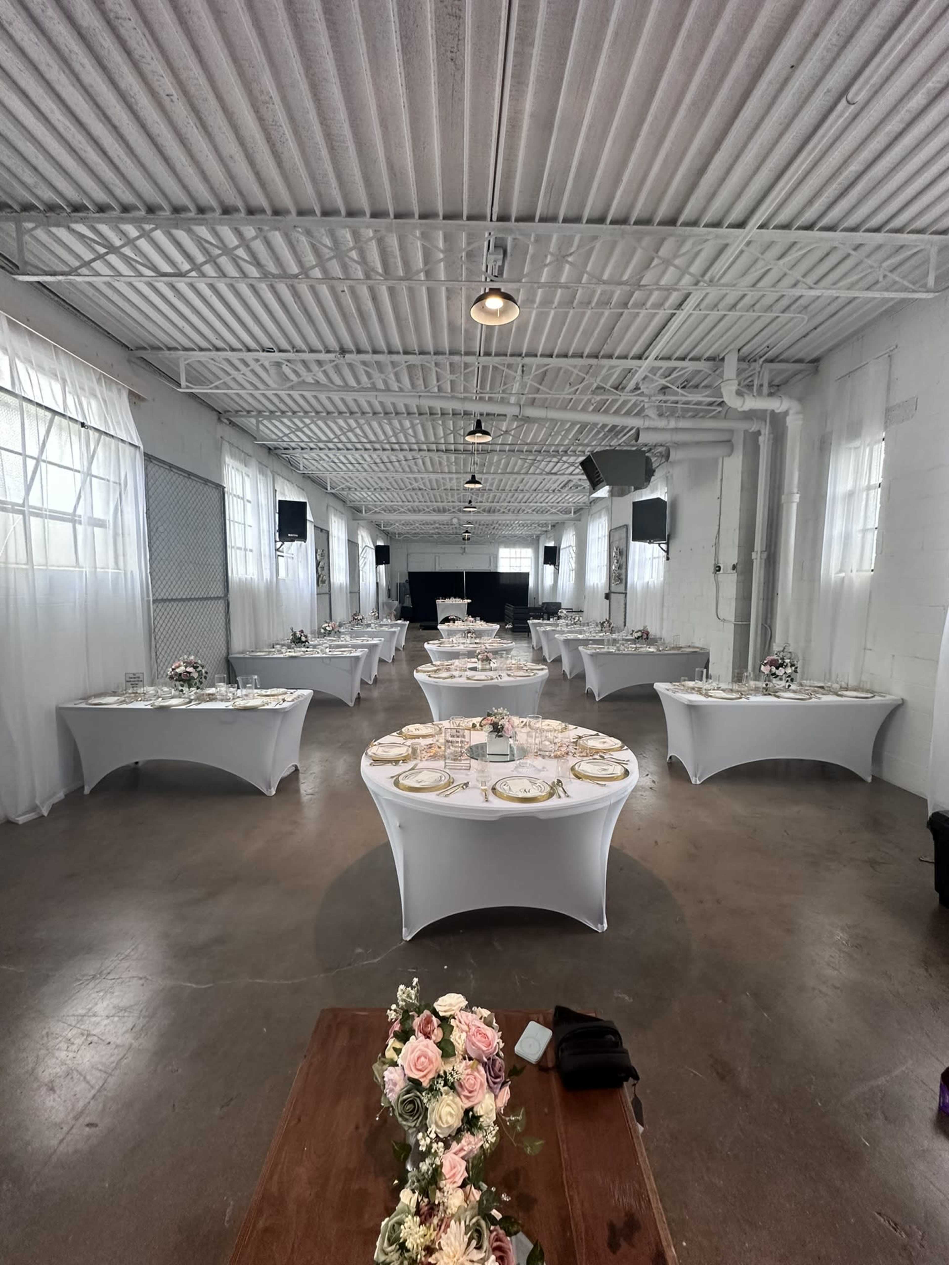 The image shows a spacious event hall set up with multiple round tables covered in white tablecloths, each arranged with diningware and floral centerpieces.