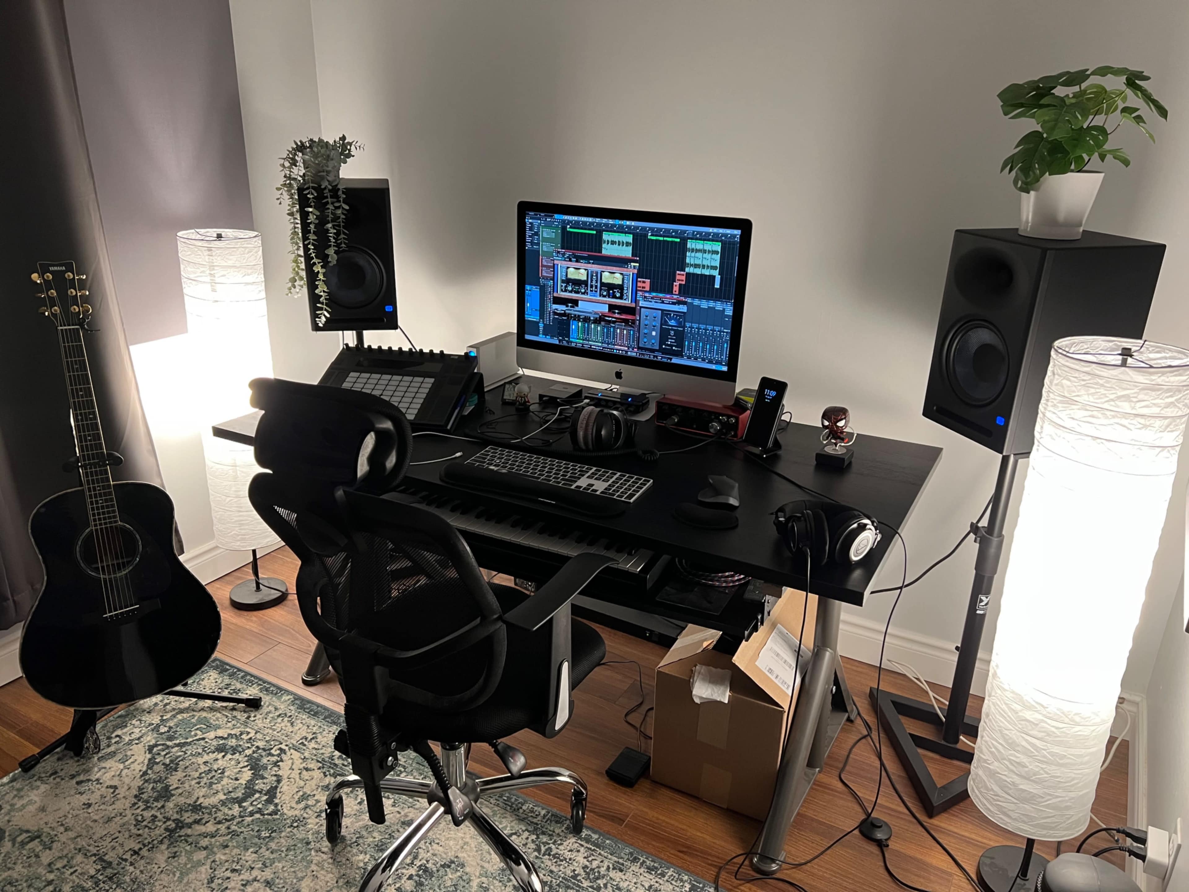A music production setup featuring a black desk with a computer, audio interface, monitors, a guitar, and ambient lighting.