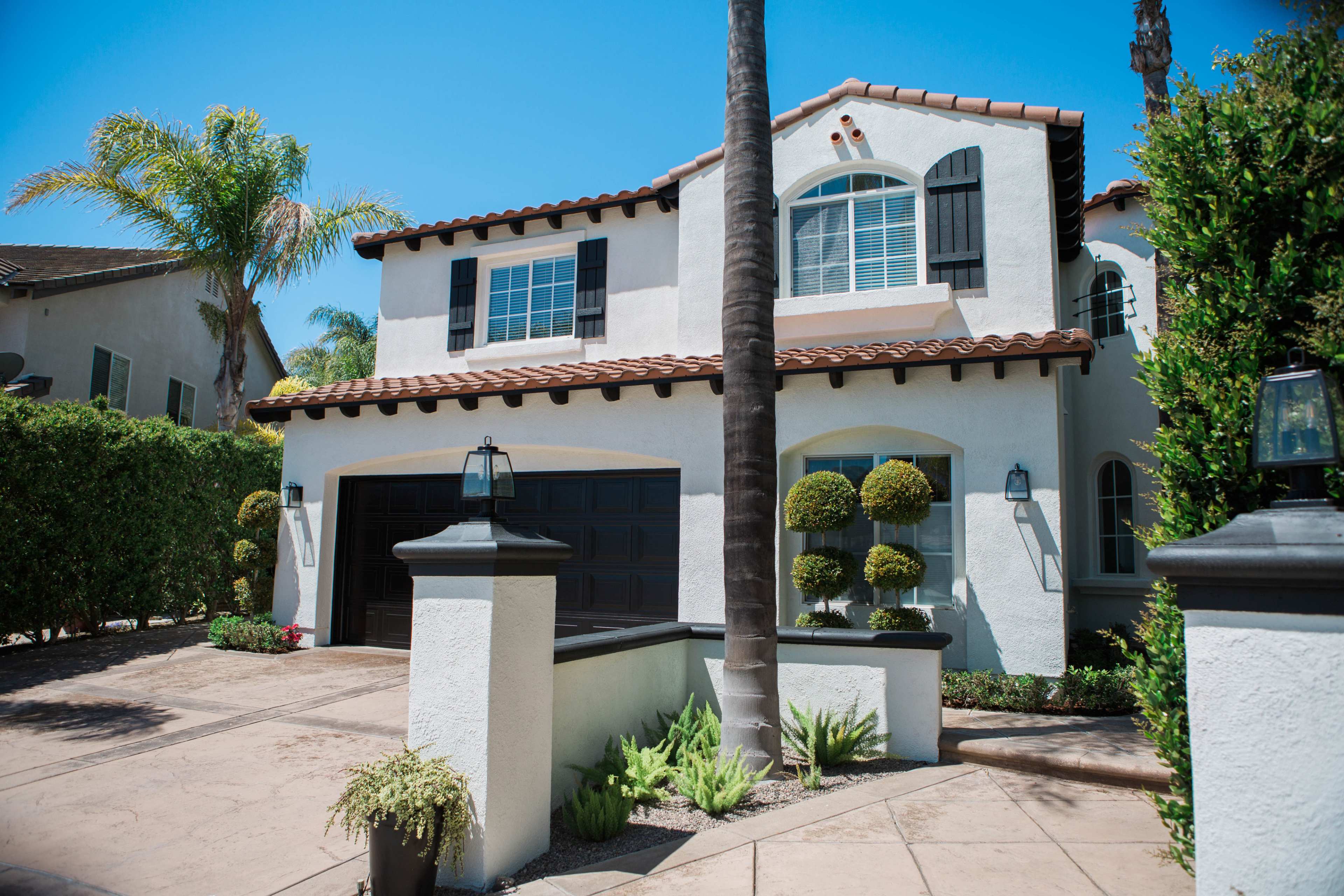 A two-story Mediterranean-style house features a tiled roof, dark shutters, and neatly trimmed hedges in the front yard.
