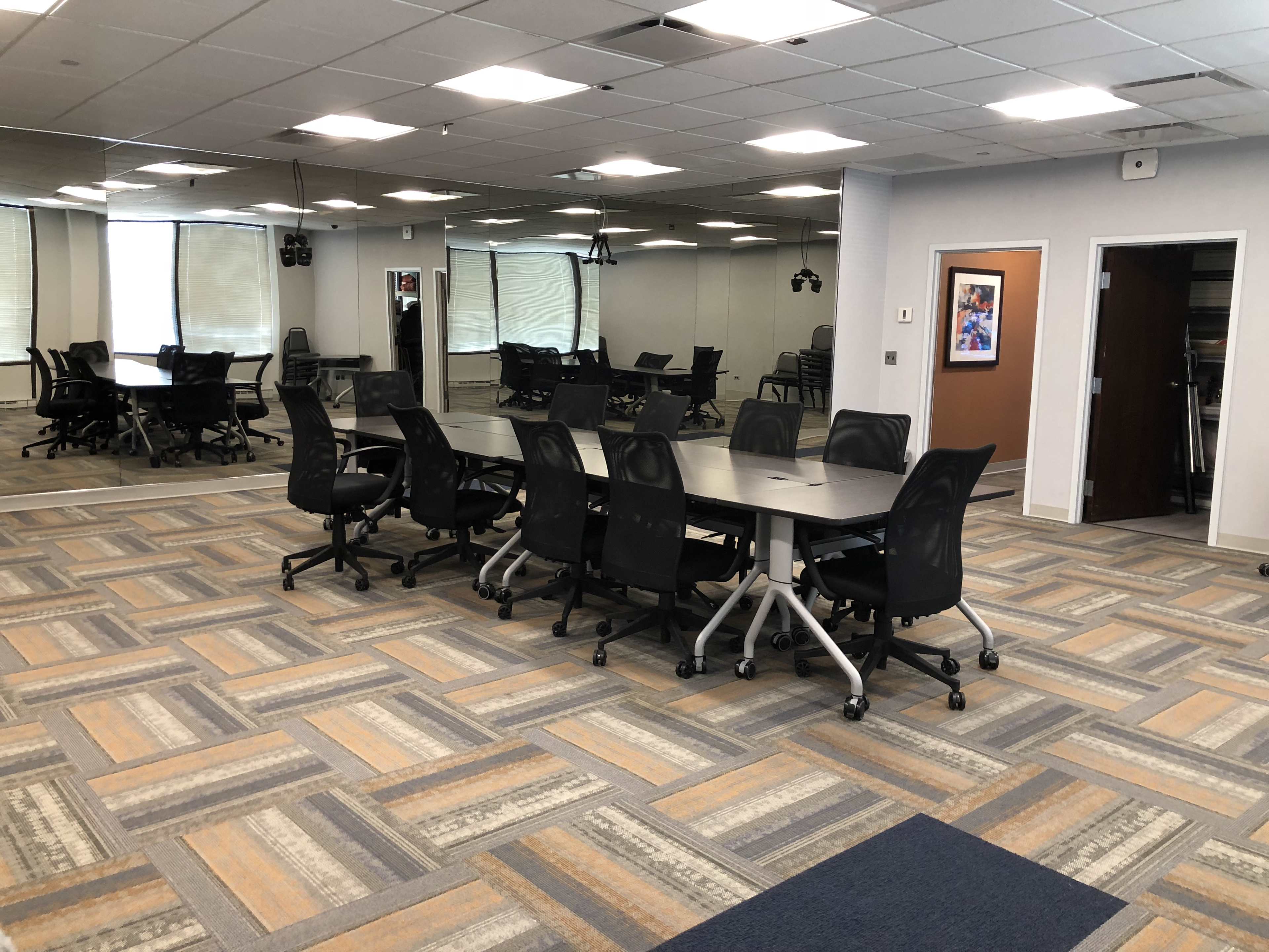 A conference room features a large table surrounded by ergonomic rolling chairs, with a mirrored wall and carpeted flooring.