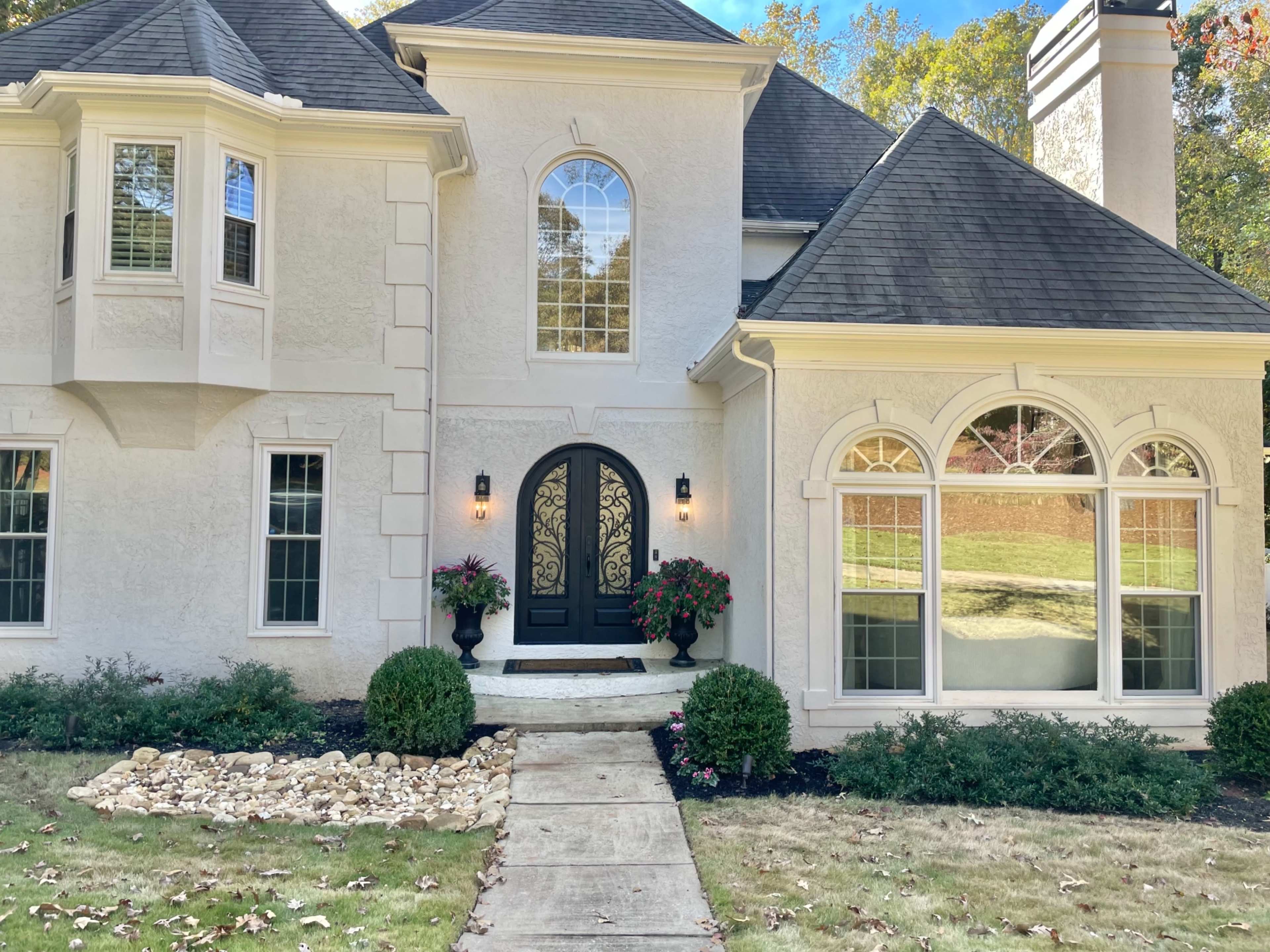 The exterior of a two-story house with a large arched front door, decorative windows, and landscaping featuring potted plants and a stone pathway.